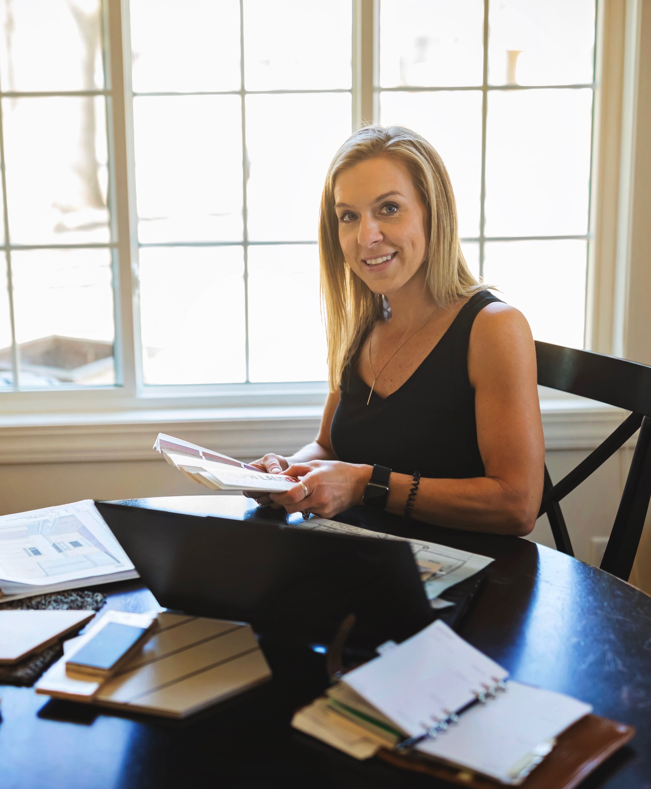 Tracy Rihn of TK Interior reviewing design materials at a table with laptop and sample boards.