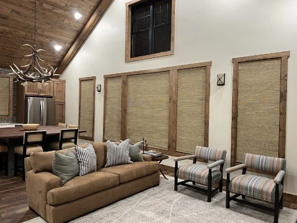 Rustic living area with wood ceiling, leather sofa, woven window shades, and seating area open to kitchen.