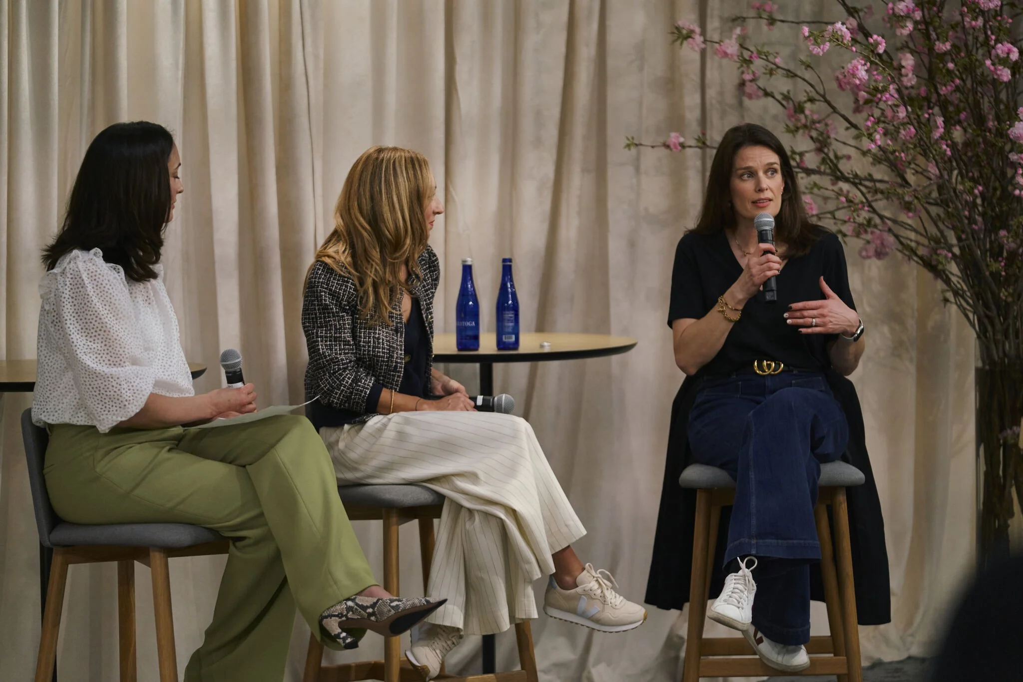 Three women engaging in a panel discussion or interview. The woman on the right is speaking into a microphone, gesturing with her left hand. The two women on the left are listening; the woman in the middle is holding a microphone, and the woman on the far left is also holding a microphone. They are seated on high stools in front of a beige curtain, with a small round table behind the middle woman that has two blue bottles on it. To the right, there is a large branch with pink blossoms.