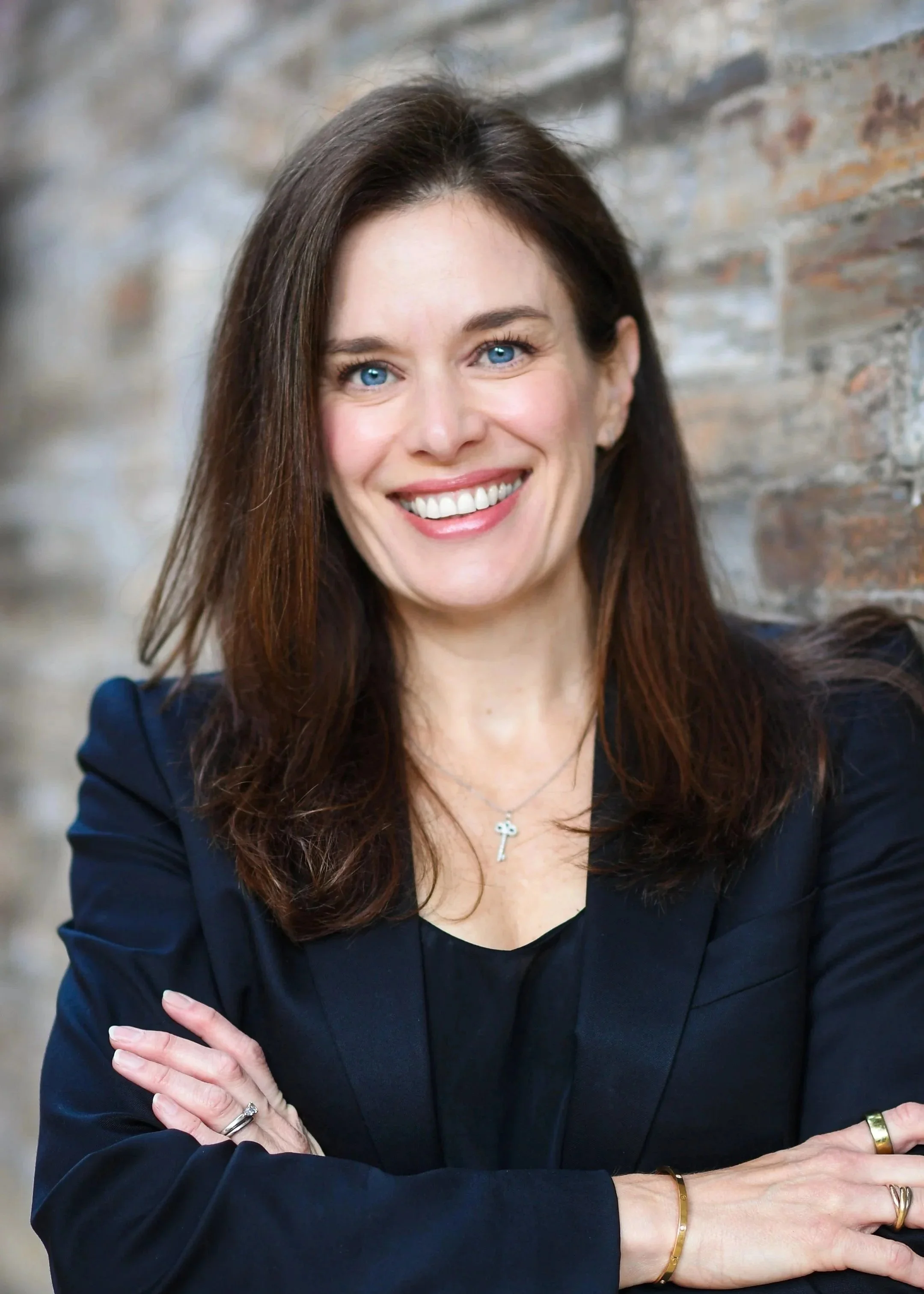 A woman with long brown hair, blue eyes, and a bright smile, wearing a black blazer and jewelry, posing with arms crossed in front of a brick wall background.