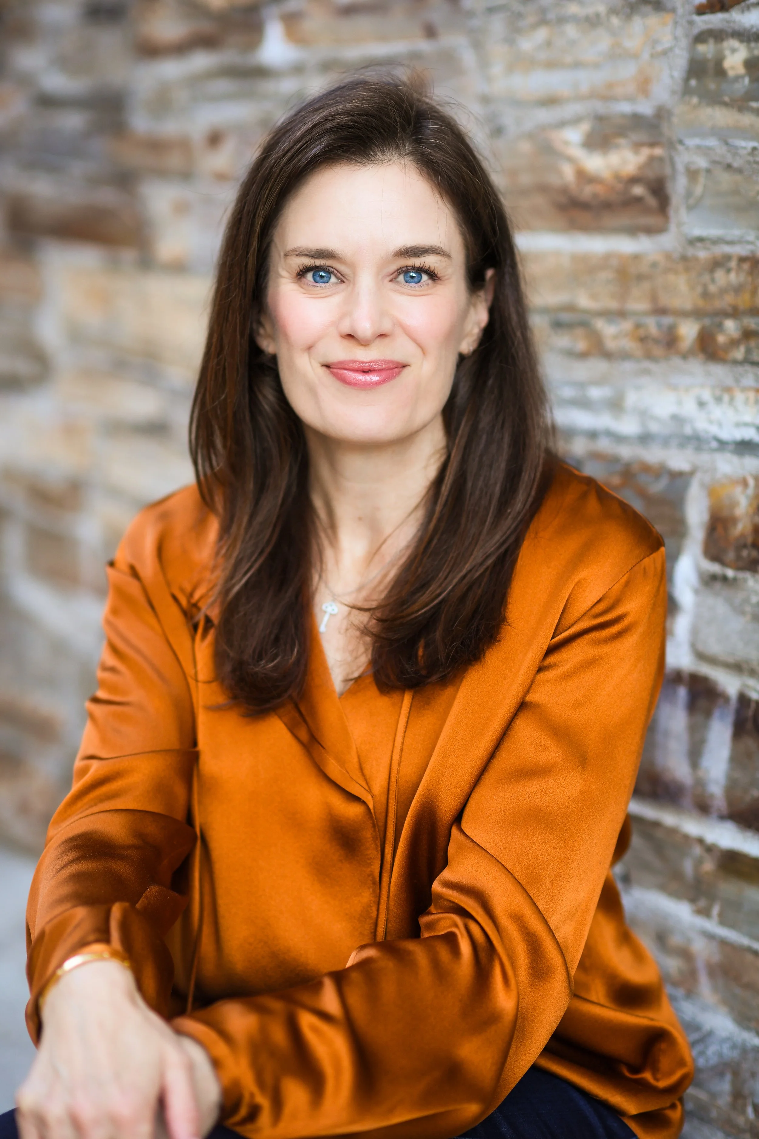 A woman with long brown hair, blue eyes, and light skin, smiling, wearing a rust-colored satin blouse, sitting against a brick wall.
