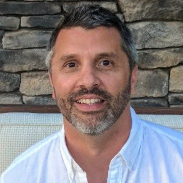 A man with short dark hair and a beard smiling, wearing a white shirt, with a stone wall background.