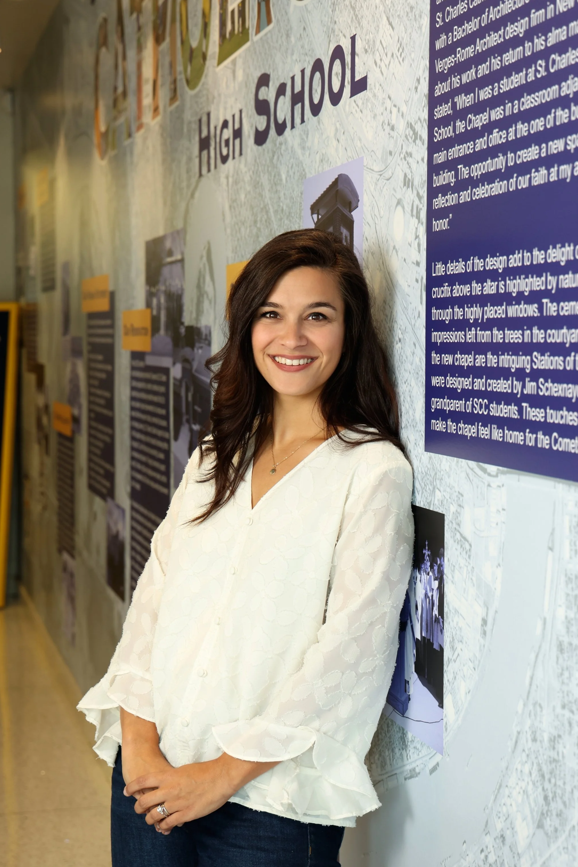 A young woman with dark brown hair and a white textured blouse smiling while standing in front of a large informational display about high school history.