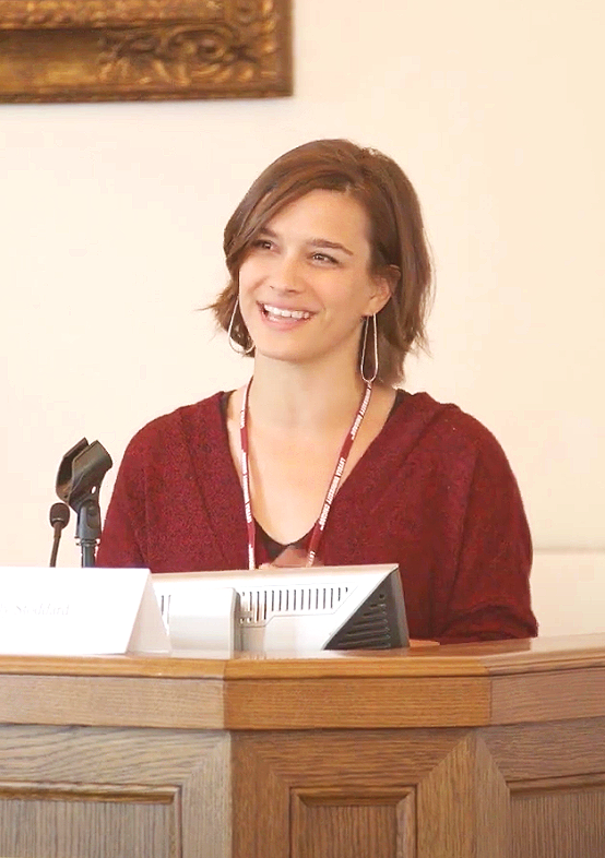 A woman with short brown hair smiling, sitting behind a wooden desk with a microphone and a computer monitor, wearing a red sweater and a lanyard, in a professional setting.