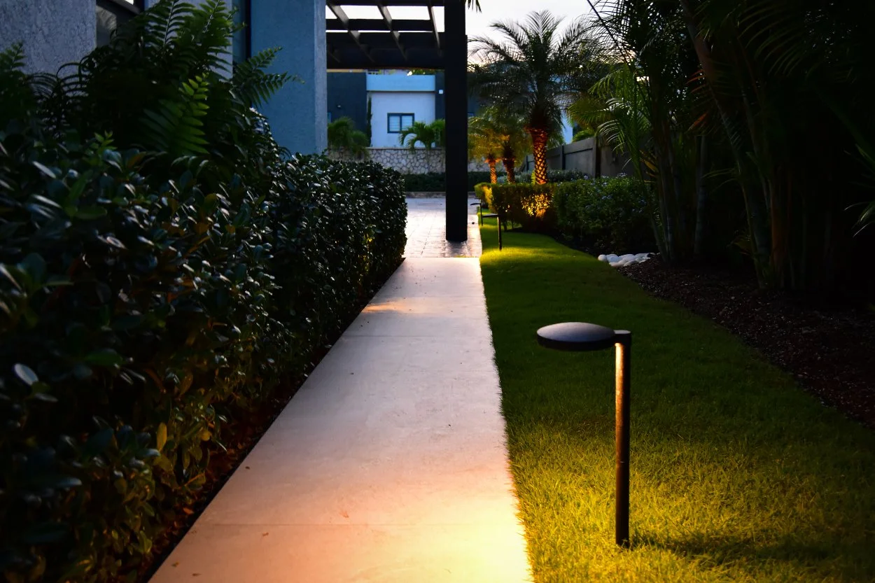 Modern path lights illuminating a garden walkway with warm lighting, highlighting hedges, grass, and palm trees in a contemporary outdoor landscape.