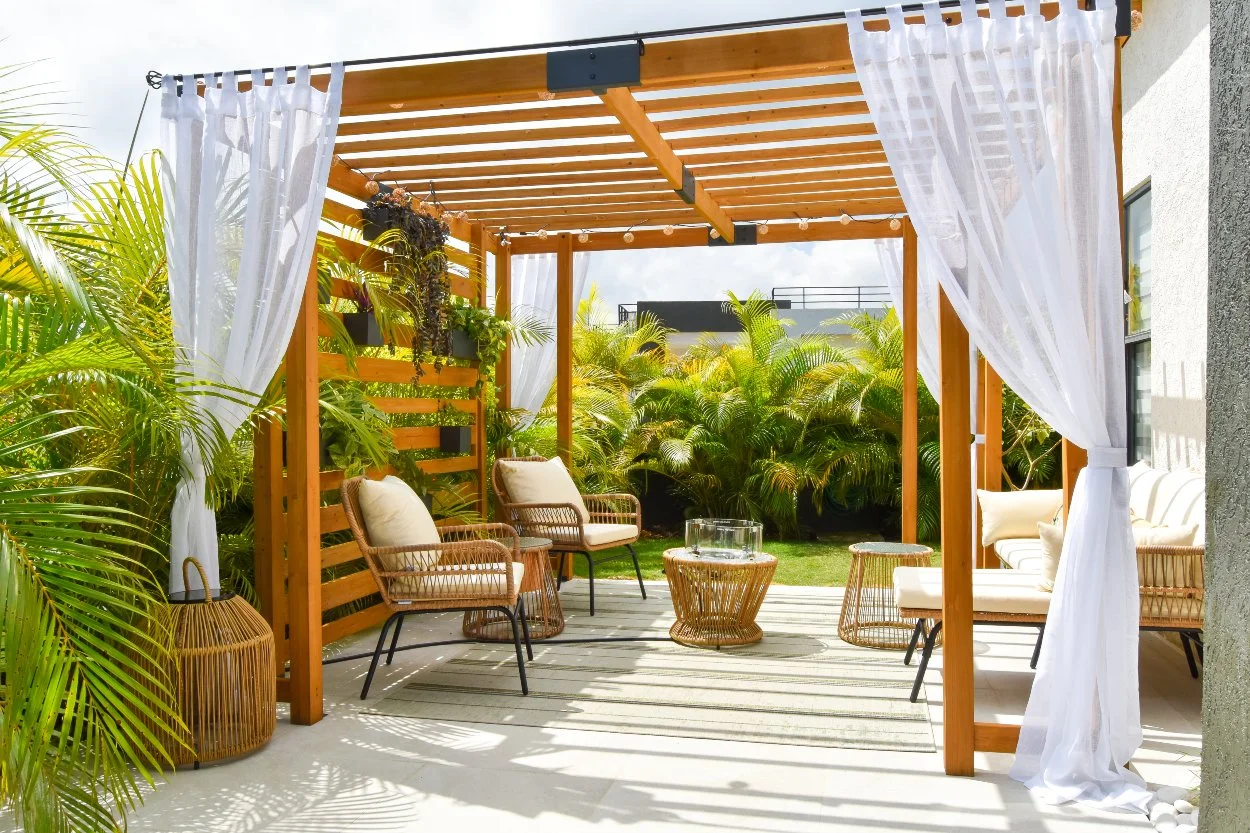 Modern backyard design featuring a wooden pergola with white drapes, wicker outdoor seating, tropical palms, and a stylish patio lounge in Jamaica.