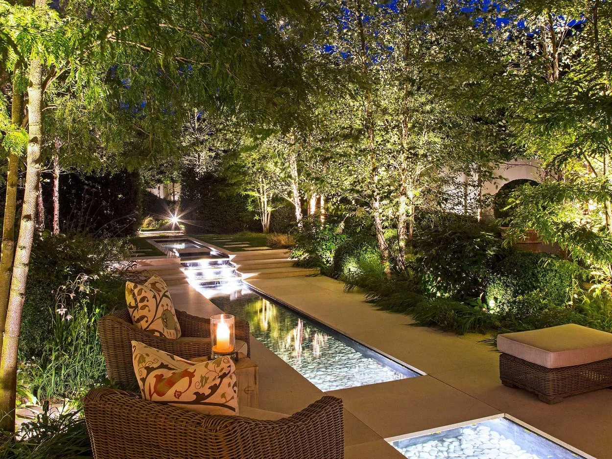 Underwater lighting illuminating a modern reflecting pool and surrounding garden, creating a glowing pathway and highlighting trees and landscape features at night.
