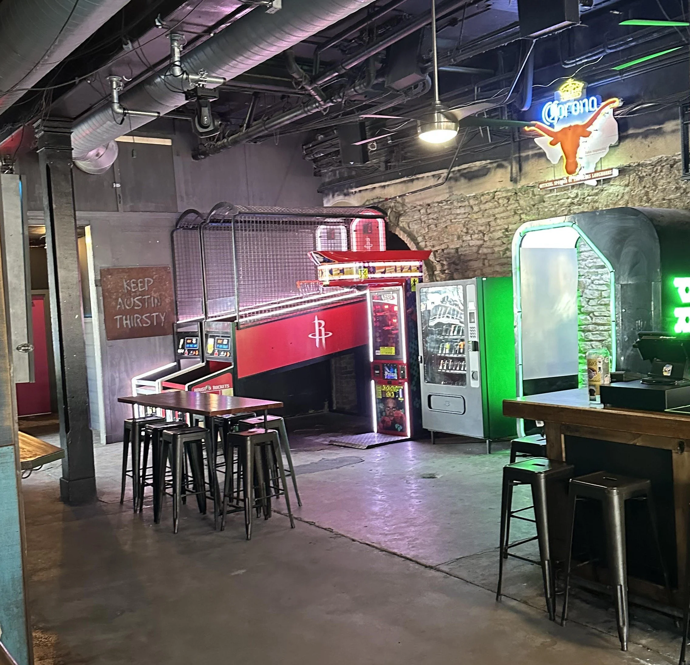 An indoor bar area with neon signs, including a Corona sign with a longhorn steer, vending machines, and arcade-style basketball game. There are tables and bar stools with colorful and industrial decor.
