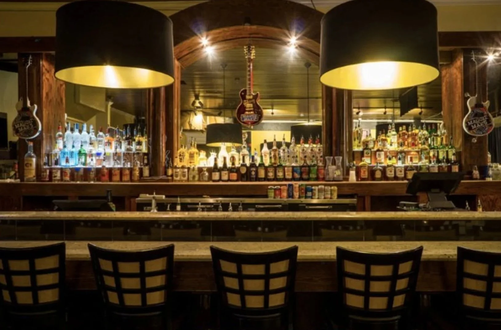 Empty bar counter with four chairs, dark hanging lamps, and shelves of bottles and alcohol in the background.