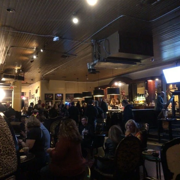A lively bar scene with people sitting and chatting at tables and gathered around a bar counter, with a bartender serving drinks. The décor includes framed pictures on the walls, a dark ceiling with ceiling lights, and a TV screen. The atmosphere is