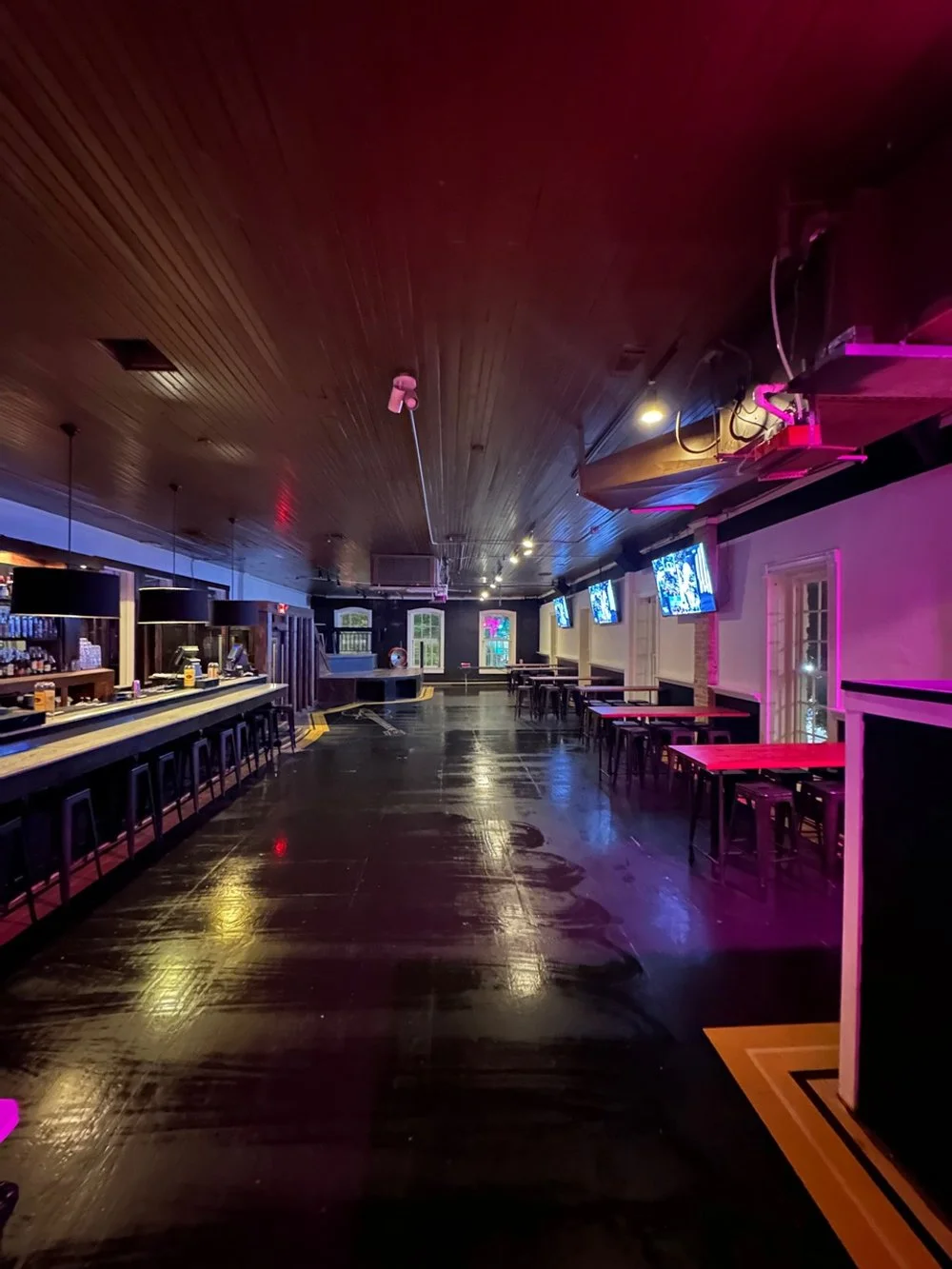 Empty bar or club interior with a long bar counter on the left, several hanging pendant lights, multiple TV screens on the right wall, and tables and chairs along the right side. Dim lighting with purple and yellow hues, wooden ceiling, and a glossy