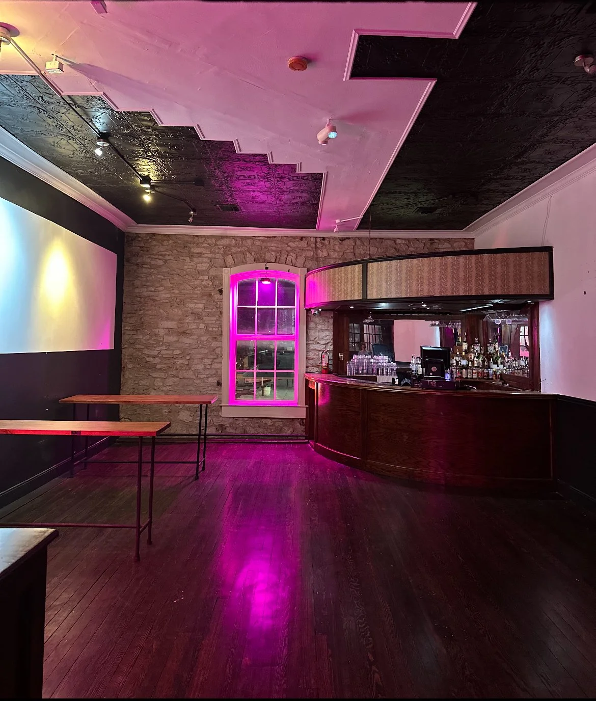 Interior of a dimly lit bar with an arched bar counter, a stone accent wall, a window with pink lighting, and empty tables with wooden tops and black metal legs.