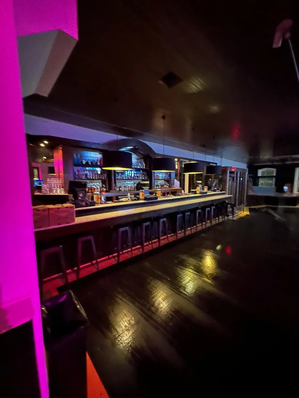 Empty bar with stools, illuminated by colorful lighting, showing bottles of drinks behind the counter in a dimly lit setting.