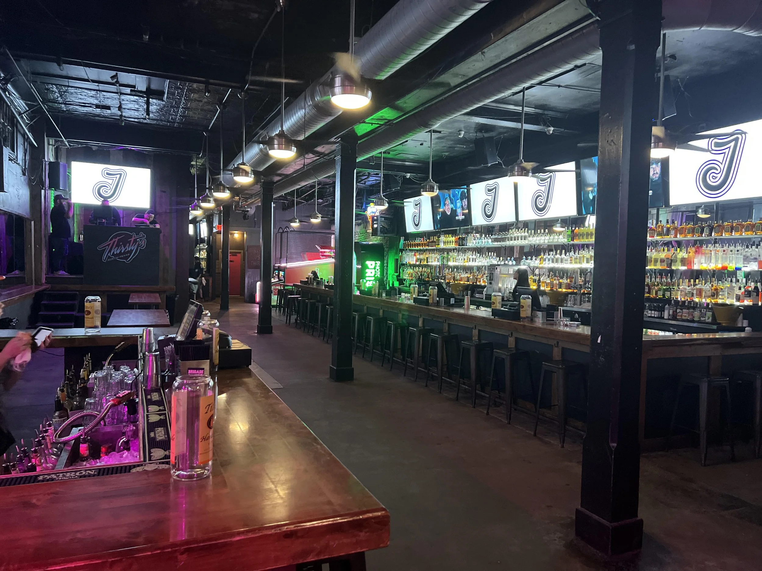 Interior of a dimly lit bar or nightclub with a long bar counter on the right filled with bottles of alcohol, high stools, and multiple overhead screens displaying a logo. There is a small bar or DJ area on the left with a sign that says "Thirsty" an