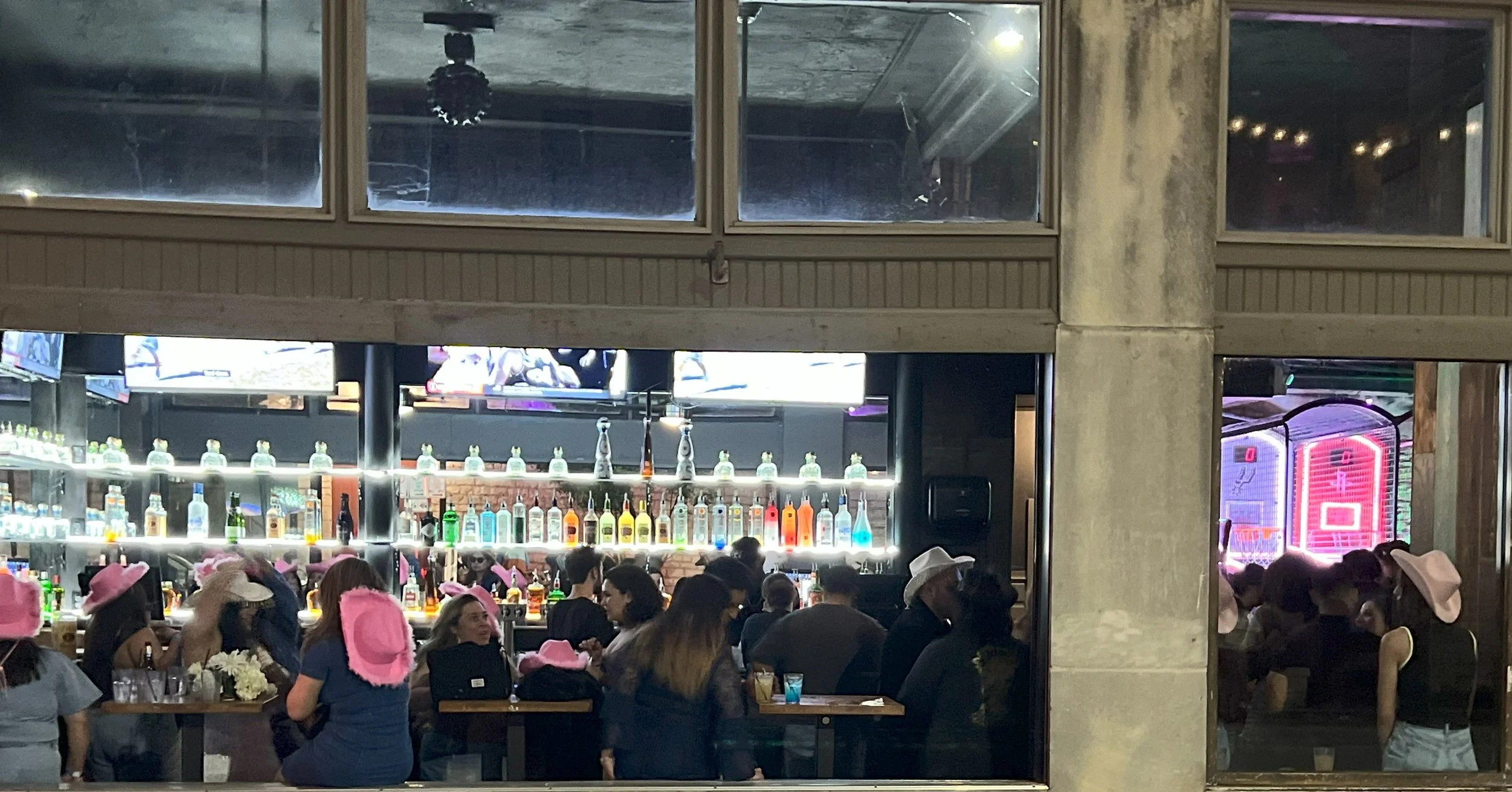 People sitting and standing inside a bar or restaurant, many wearing pink hats. The bar has shelves with bottles and neon lights, and television screens mounted on the wall.