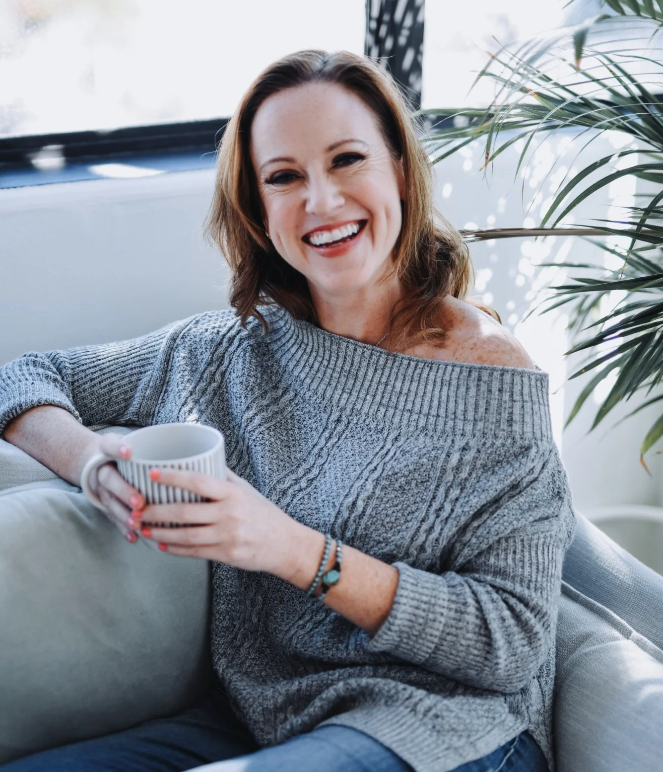 A woman with brown hair smiling and holding a white mug, sitting on a couch near a window with sunlight and green plants in the background.