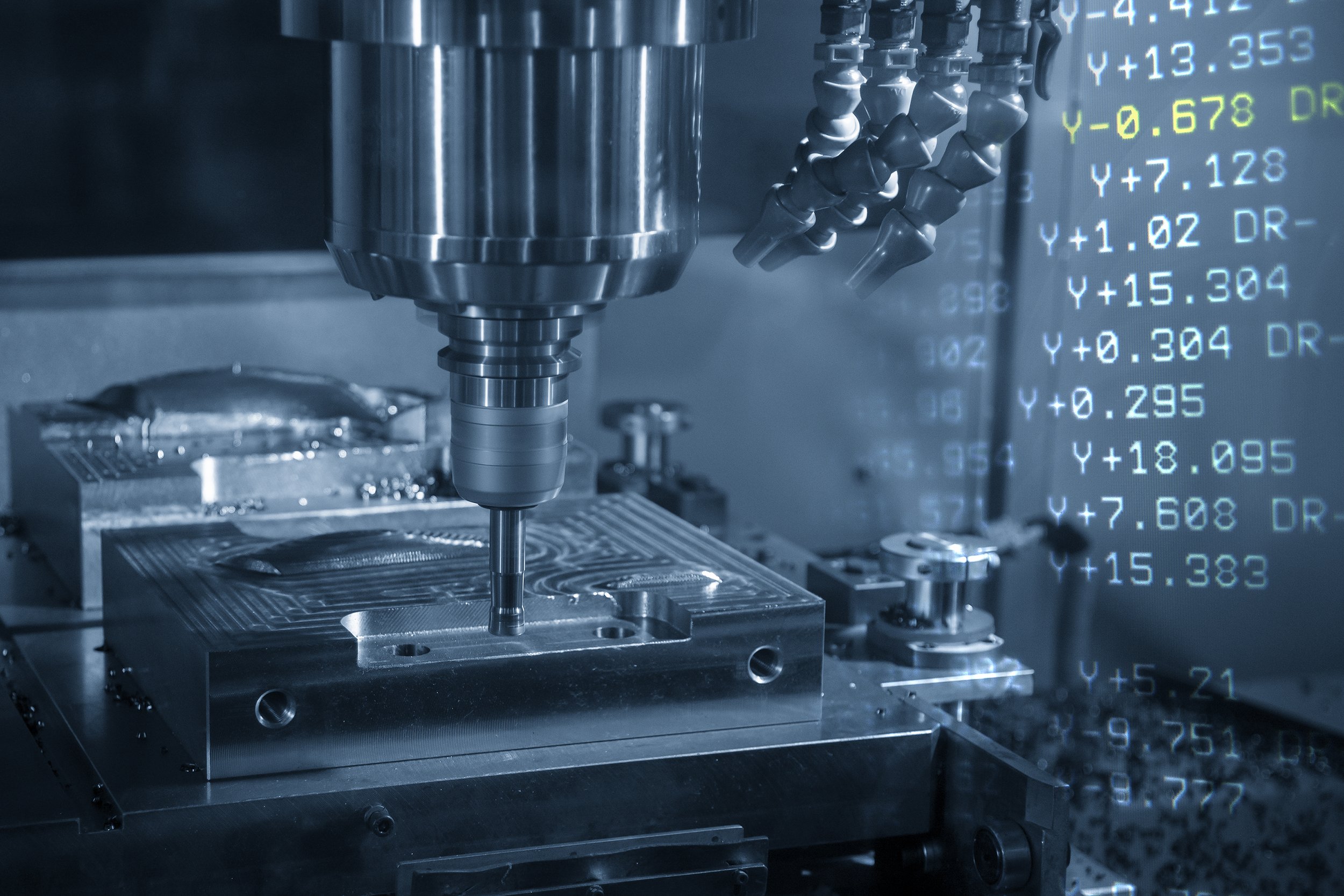 Close-up of a CNC milling machine with a drill bit working on a metal workpiece, with digital readouts visible on a screen in the background.