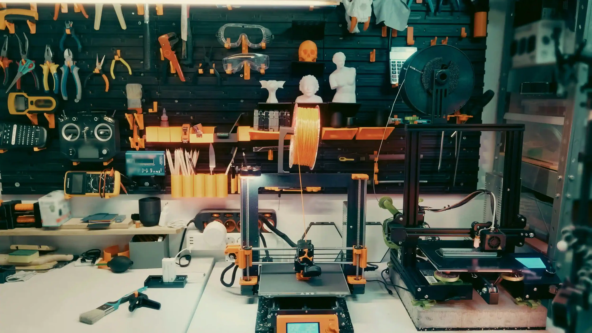 A workspace with 3D printers, tools, and art sculptures, including busts and skulls, on a black pegboard wall.