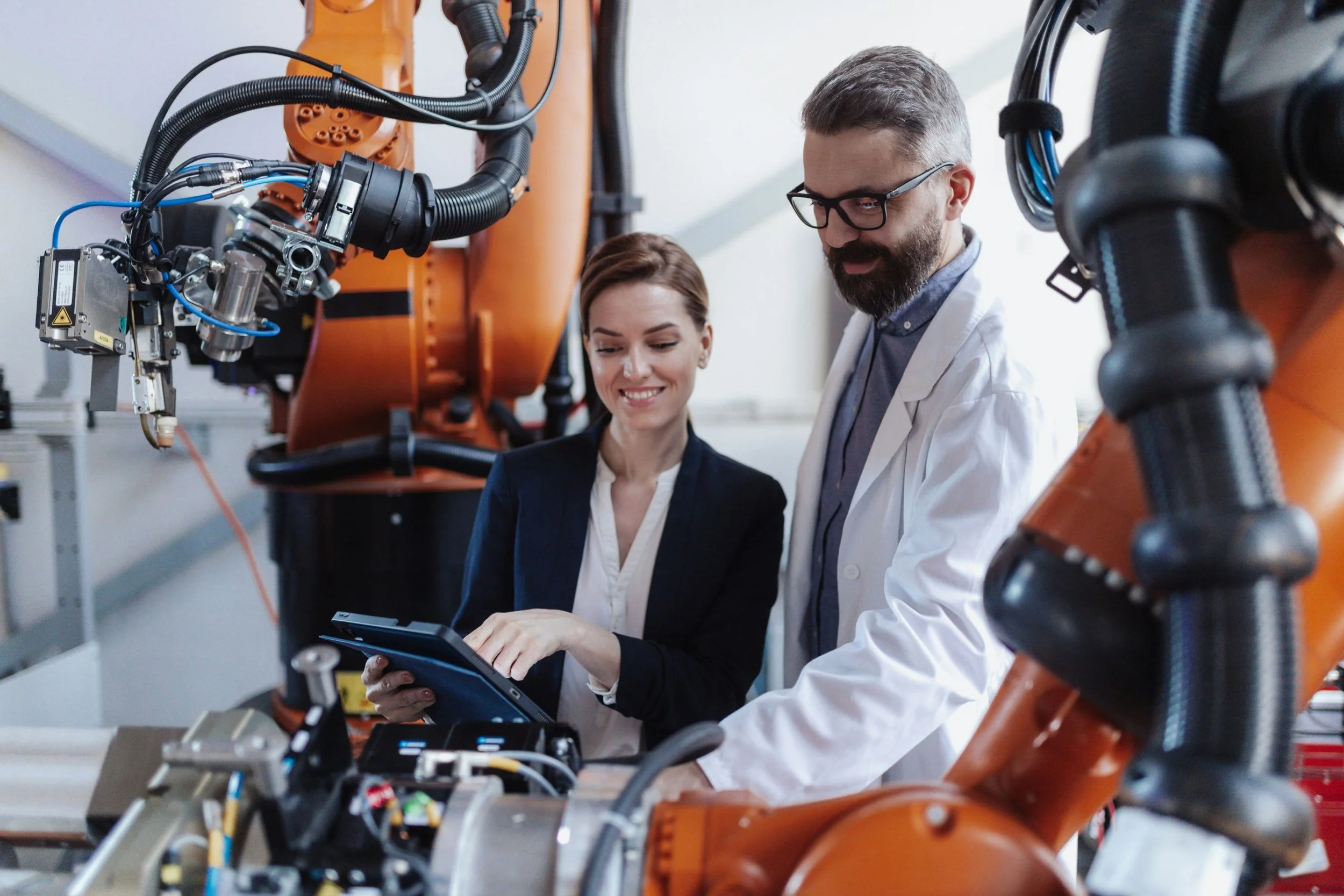 Three people, two men and one woman, working with industrial robotic arms in a laboratory or factory setting. The woman is holding a tablet and smiling, while the man in a white lab coat is observing and smiling. The robotic arms are large and orange, connected with various cables and equipment.