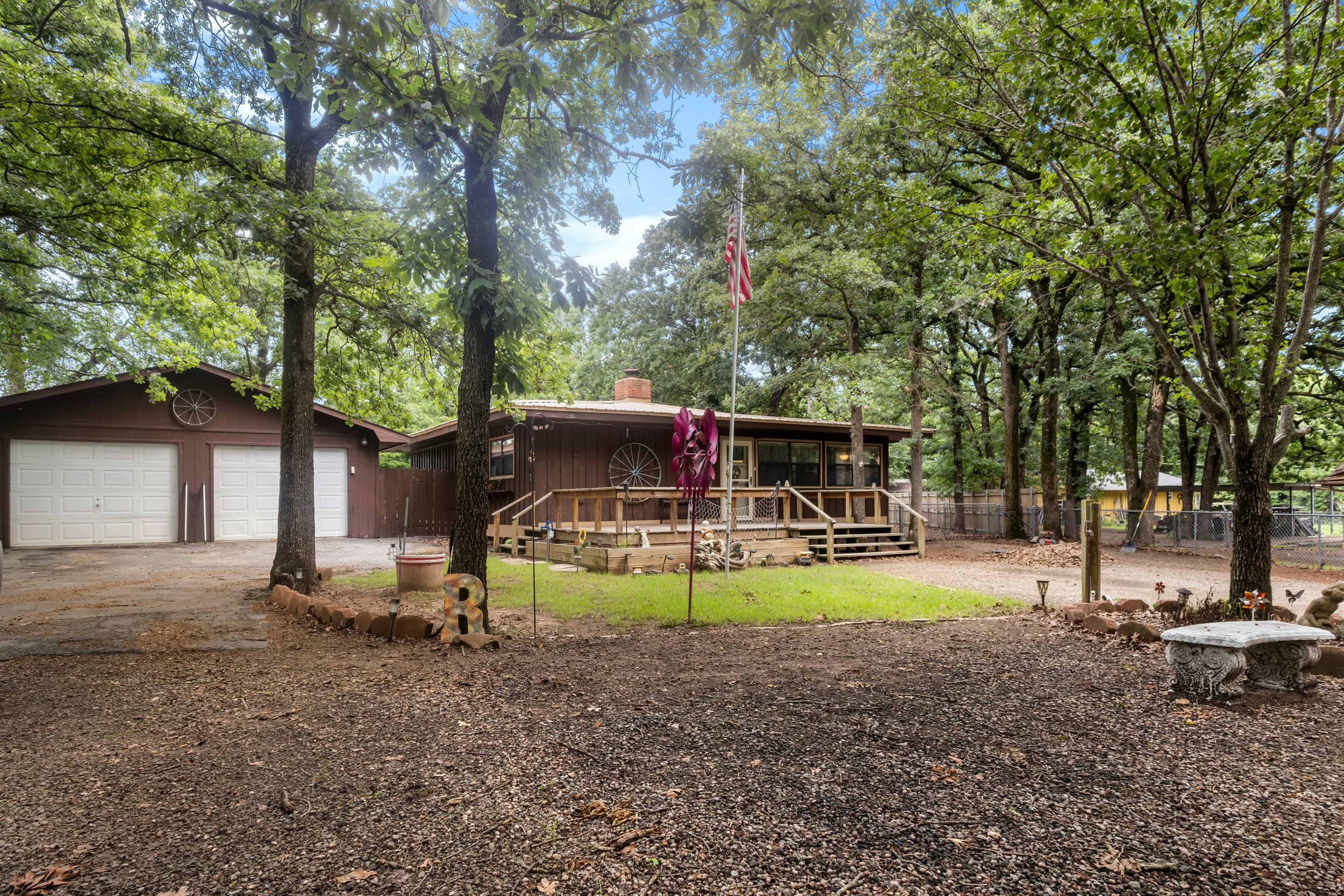 Backyard with large trees, a brown house with a deck, American flags, decorative windmills, a stone birdbath, and a garden area with small plants and garden ornaments.