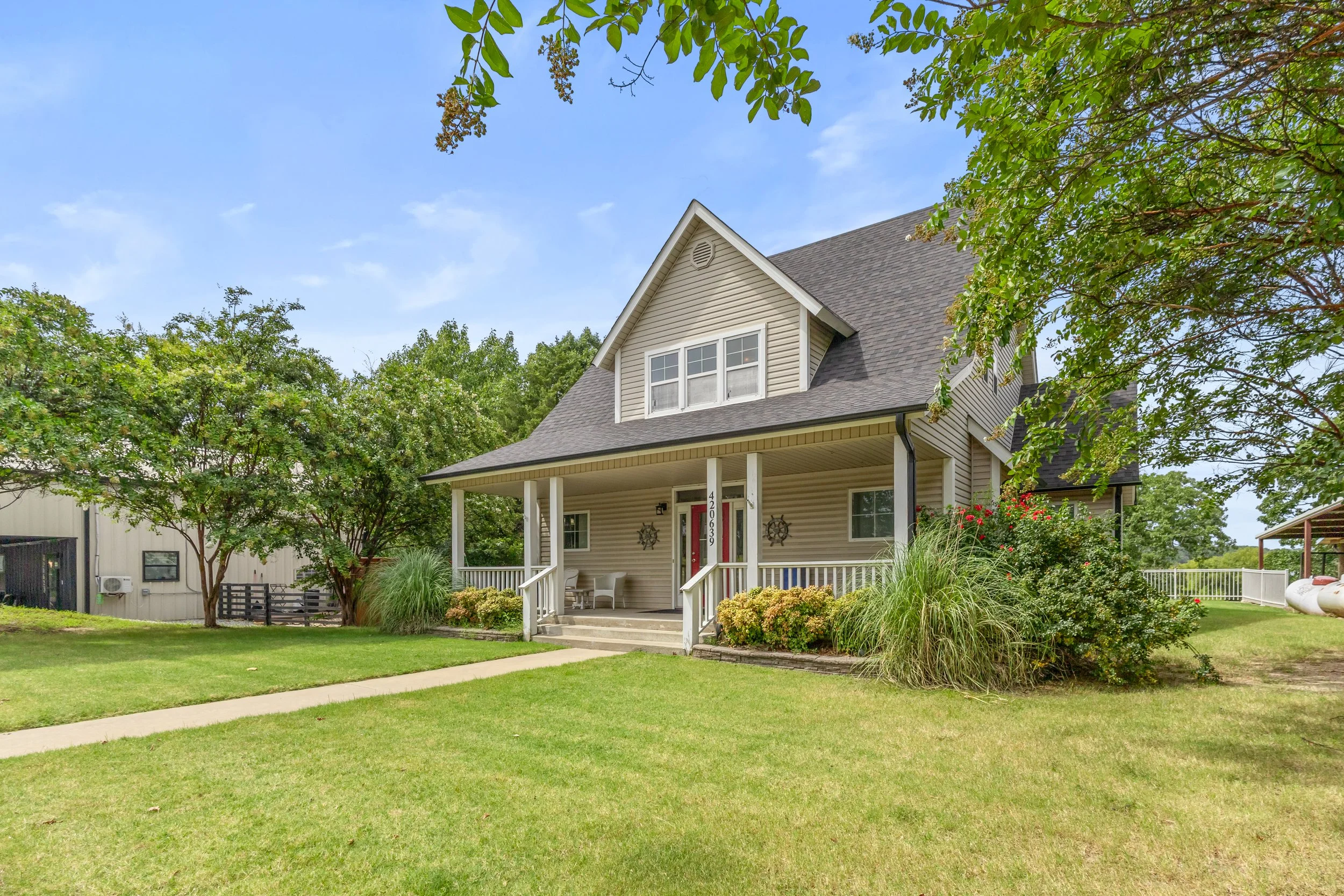 A two-story house with beige siding and a gabled roof surrounded by green trees and lawn, with a porch and seating area.