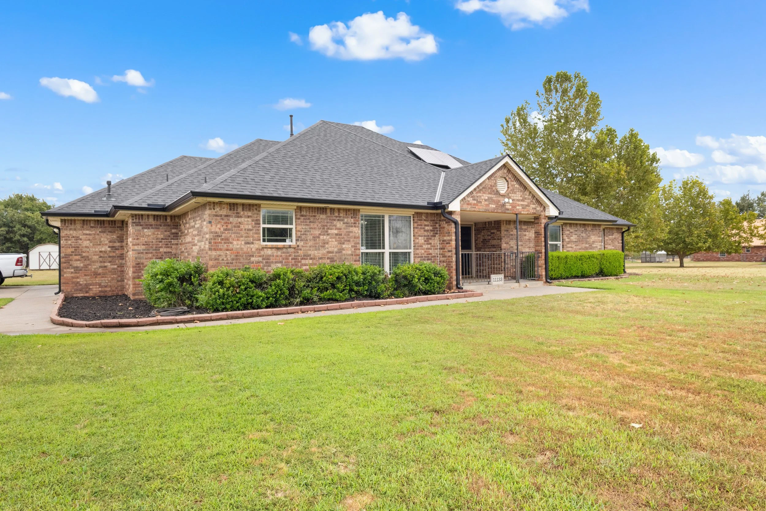 Front view of a brick house with a gray shingled roof, solar panels, landscaped bushes, and a green lawn under a blue sky with scattered clouds.