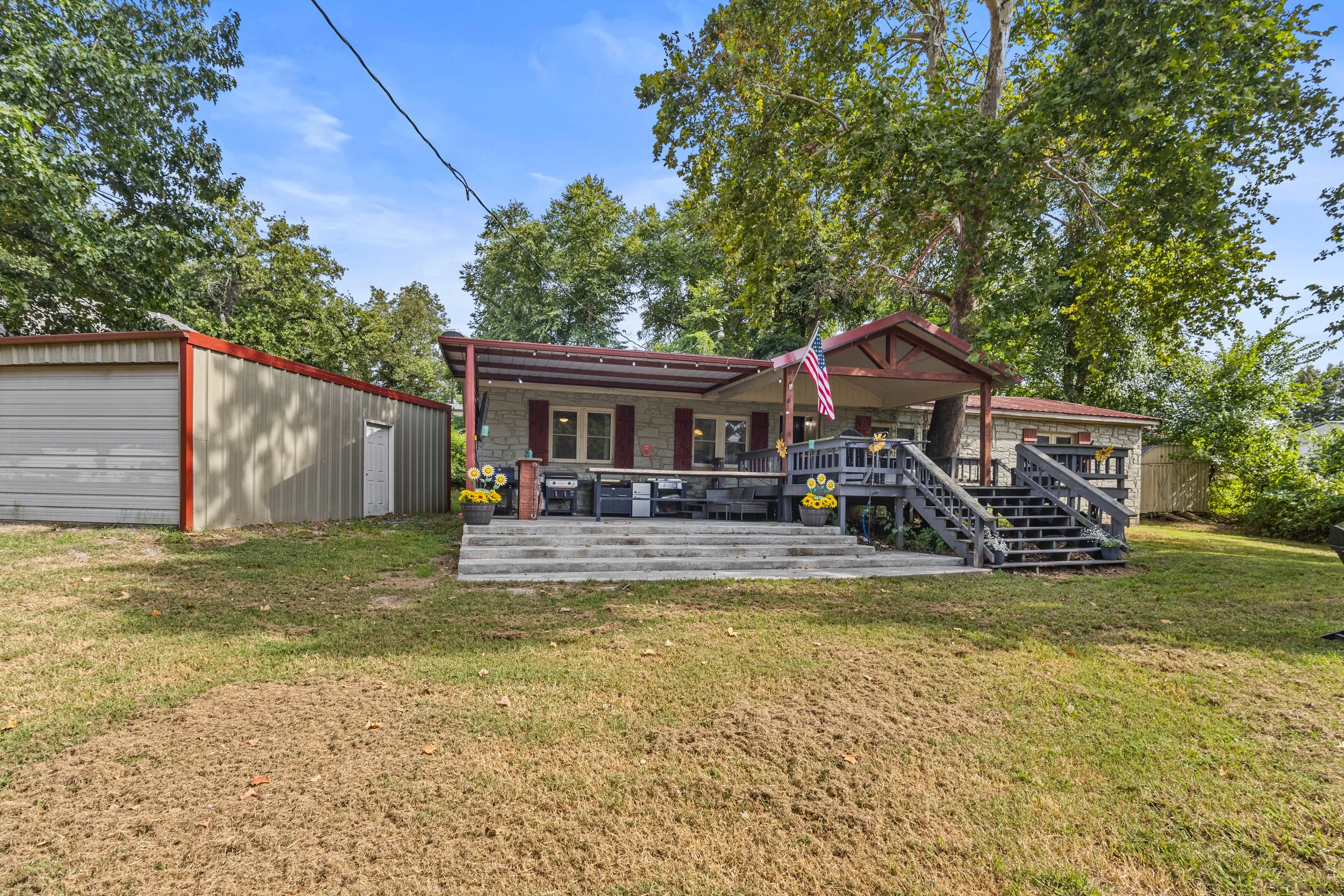 A house with a porch decorated with American flags, surrounded by lush green trees, with a metal storage shed to the left, and a lawn area in the foreground.