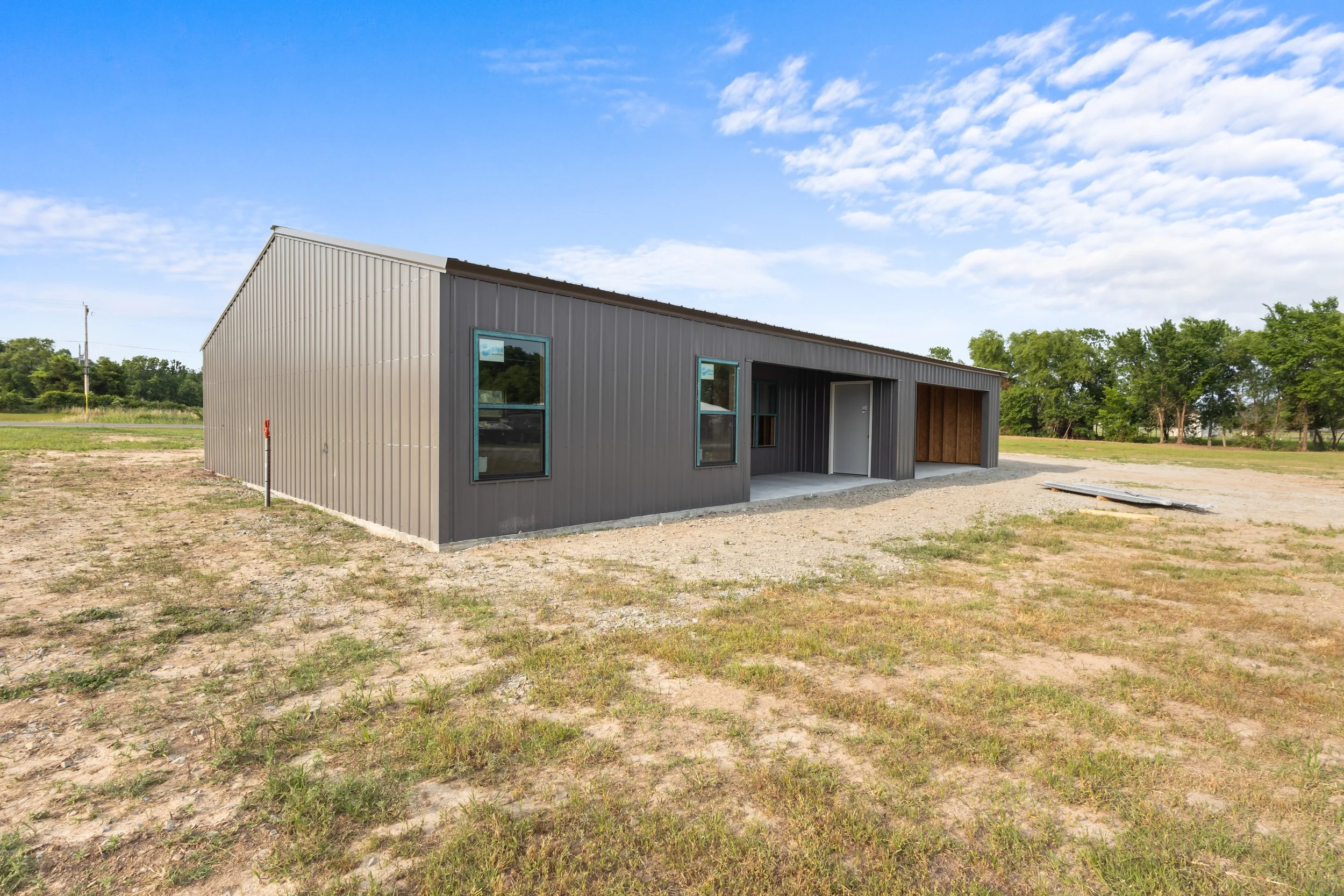 A metal building with a sloped roof under a partly cloudy sky, surrounded by a grassy and dirt area, with some trees in the background.