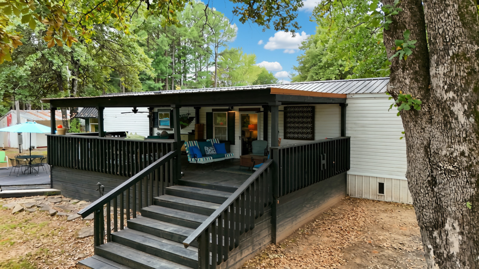 Back porch of a house with black railing and stairs, decorated with outdoor furniture including a swing, pillows, and chairs. Trees and a blue sky with clouds in the background.