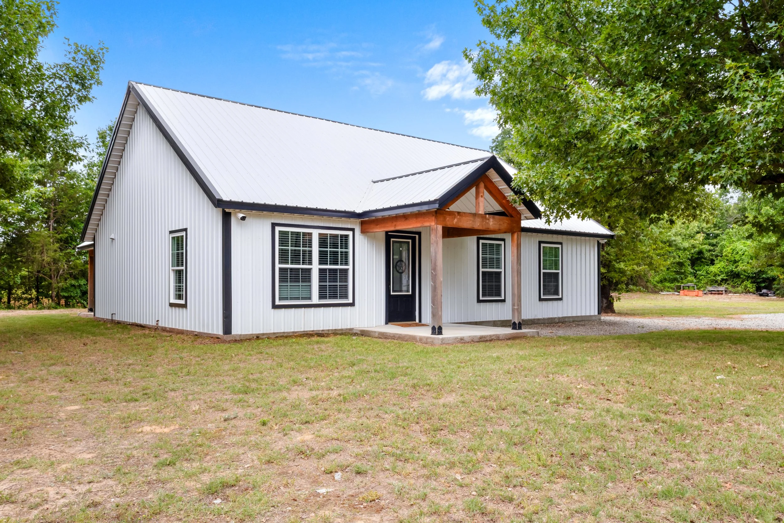 White house with black trim and a metal roof, surrounded by green trees and grass.
