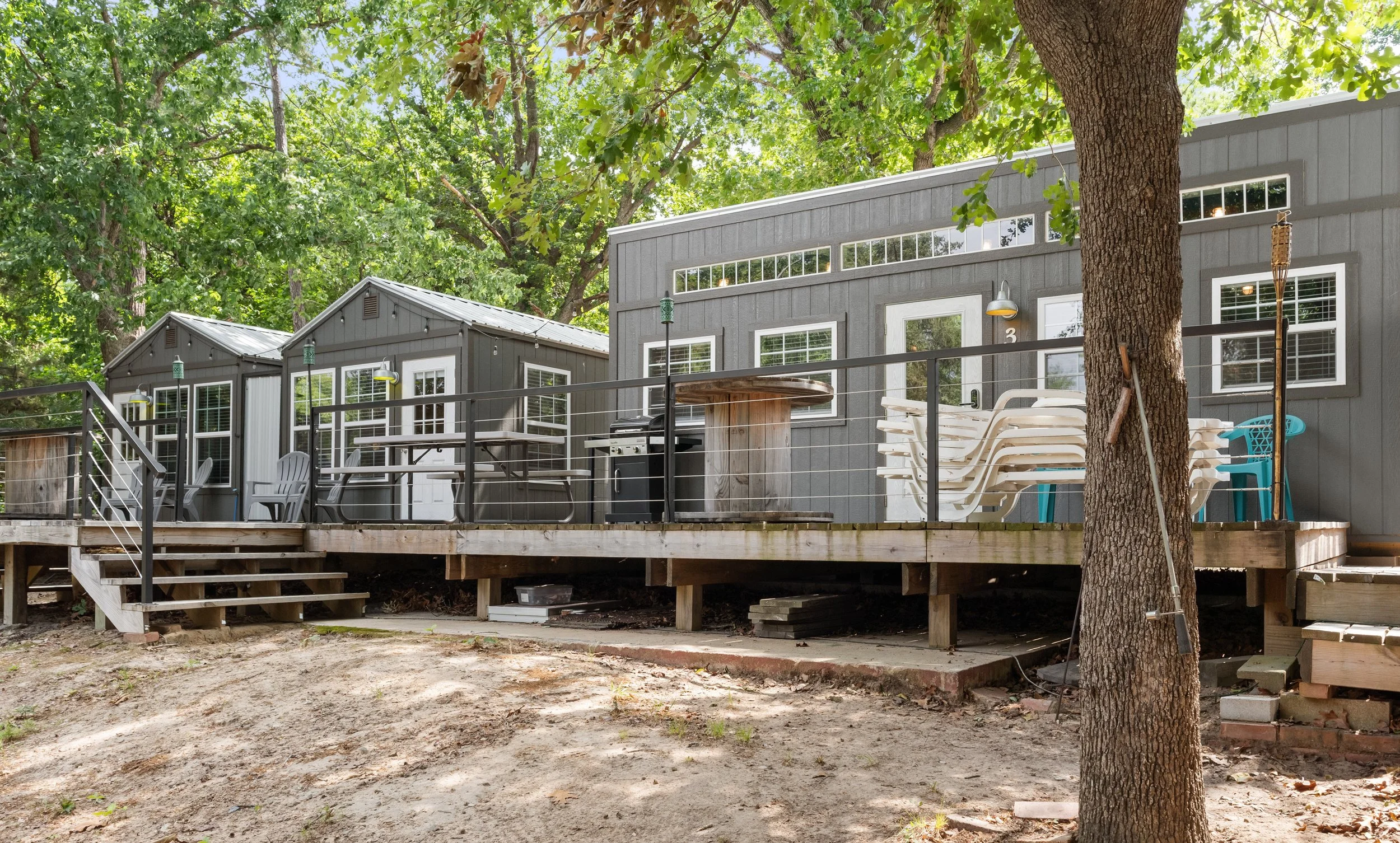 A wooden deck attached to a gray house with multiple windows, surrounded by green trees, with outdoor chairs, a barbecue grill, and a stack of plastic chairs. The deck has stairs leading down to the ground.