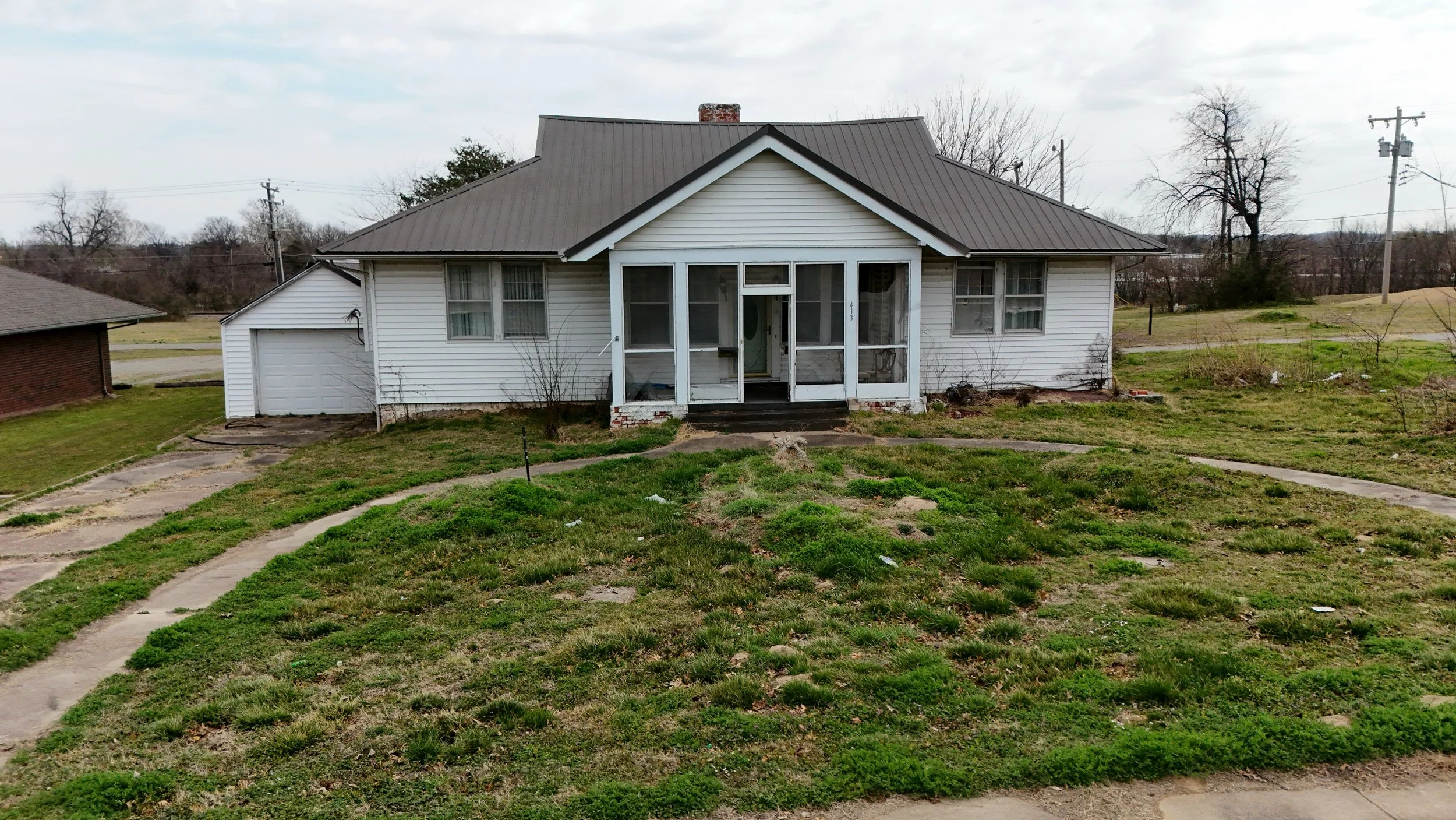 A detached white house with a metal roof, front porch steps, and a lawn with patchy grass and concrete walkways.