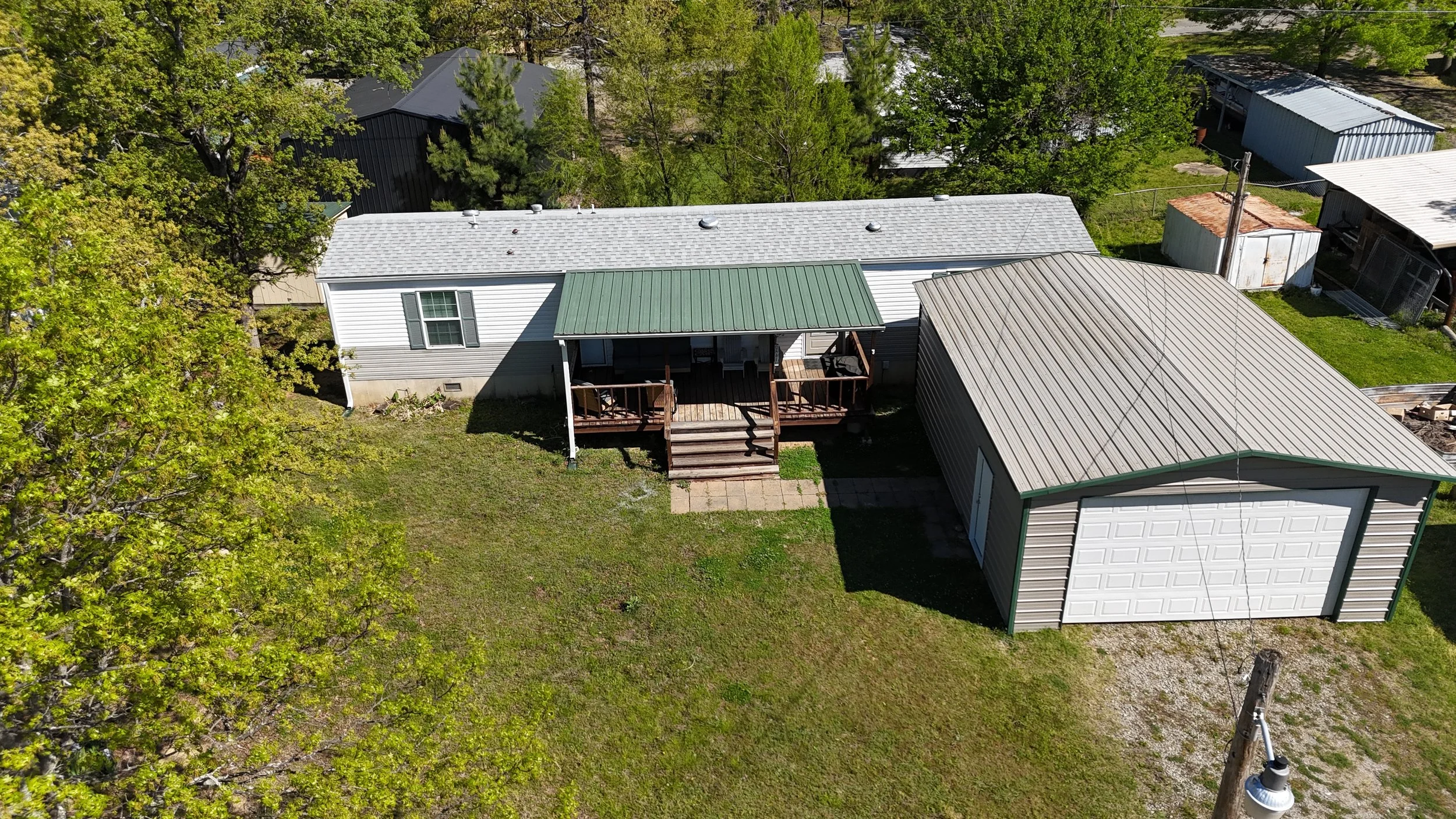 Aerial view of a house with a backyard deck, surrounded by green trees and neighboring buildings, with a driveway and utility pole.