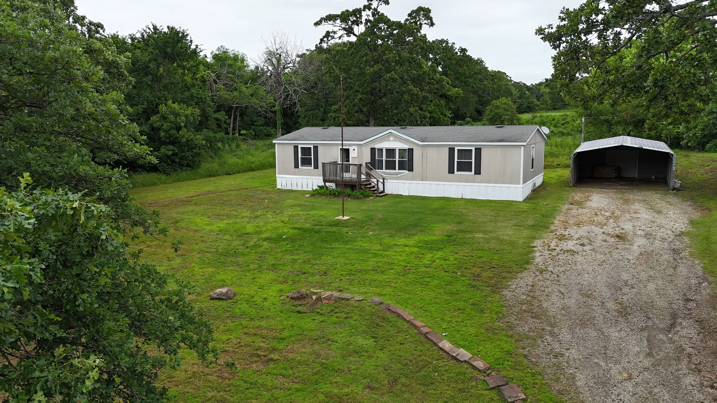 A single-story mobile home with grey siding, black shutters, and a small wooden deck at the front entrance. There is a gravel driveway leading to a covered carport on the right. Surrounding the house is a grassy yard with some trees and bushes, with a wooded area in the background.