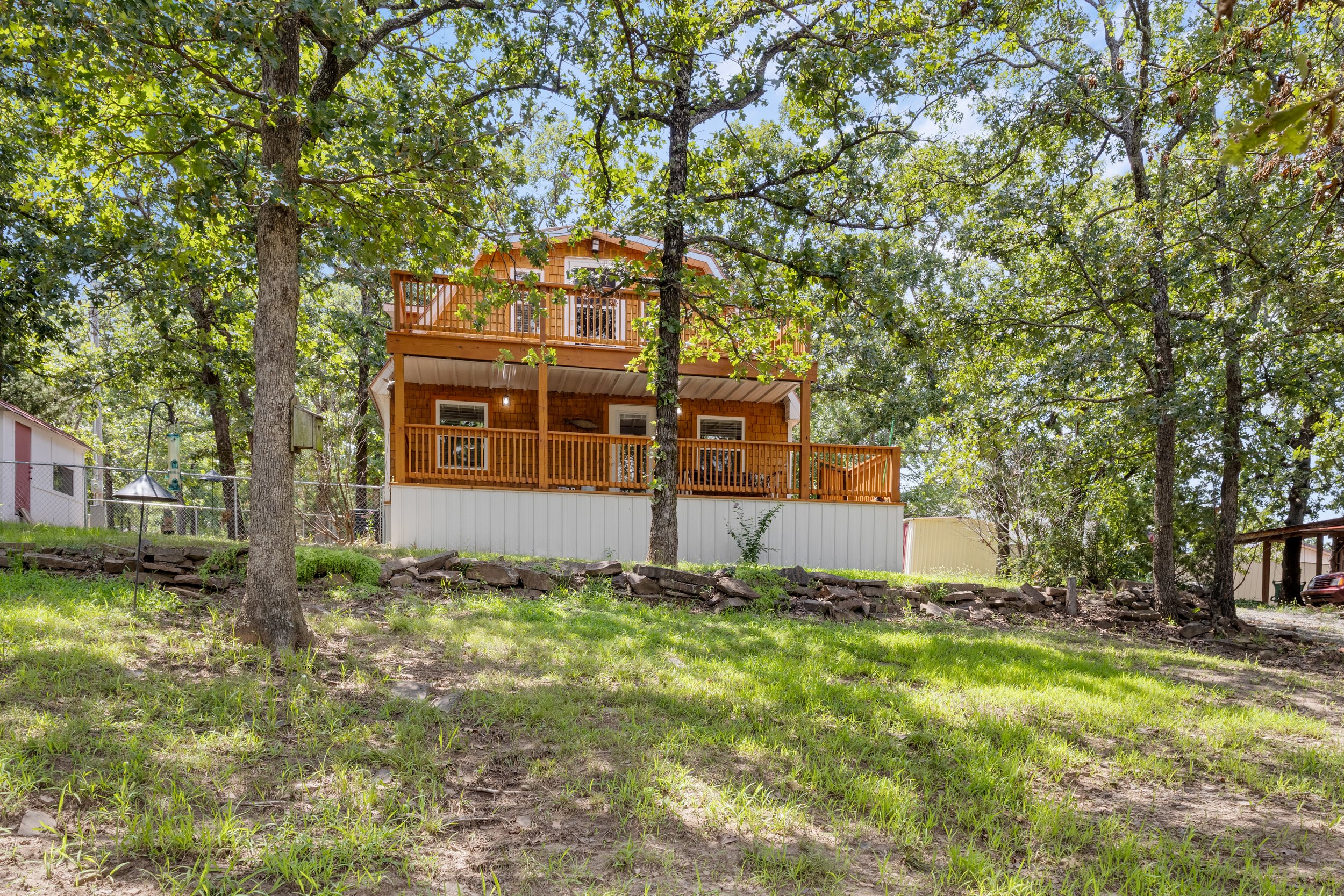 A house with a large wooden deck is surrounded by tall trees with green leaves, on a sunny day in a backyard with grass and rocks.
