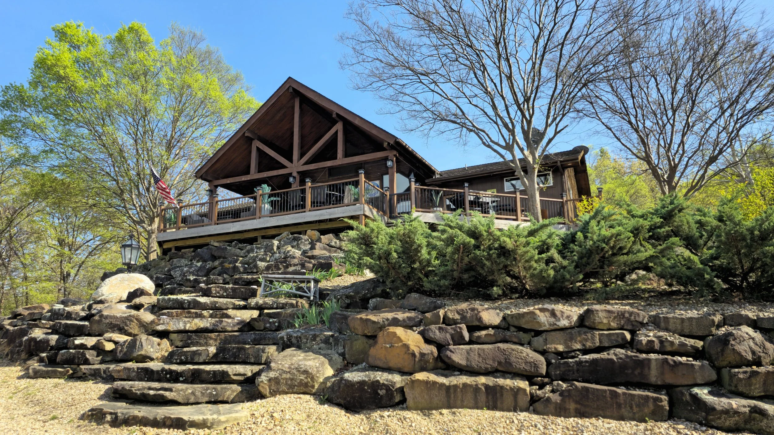 A wooden house on a hill with a stone retaining wall and a deck, surrounded by trees with some bare branches and some green leaves, under a blue sky.