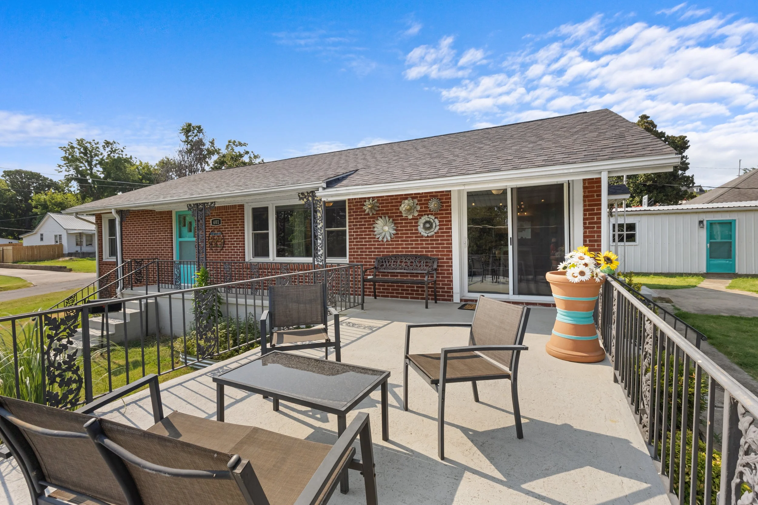Front porch of a brick house with a guesthouse in the background, decorated with metal wall art and a large flower pot with daisies, overlooking a well-maintained yard under a partly cloudy sky.