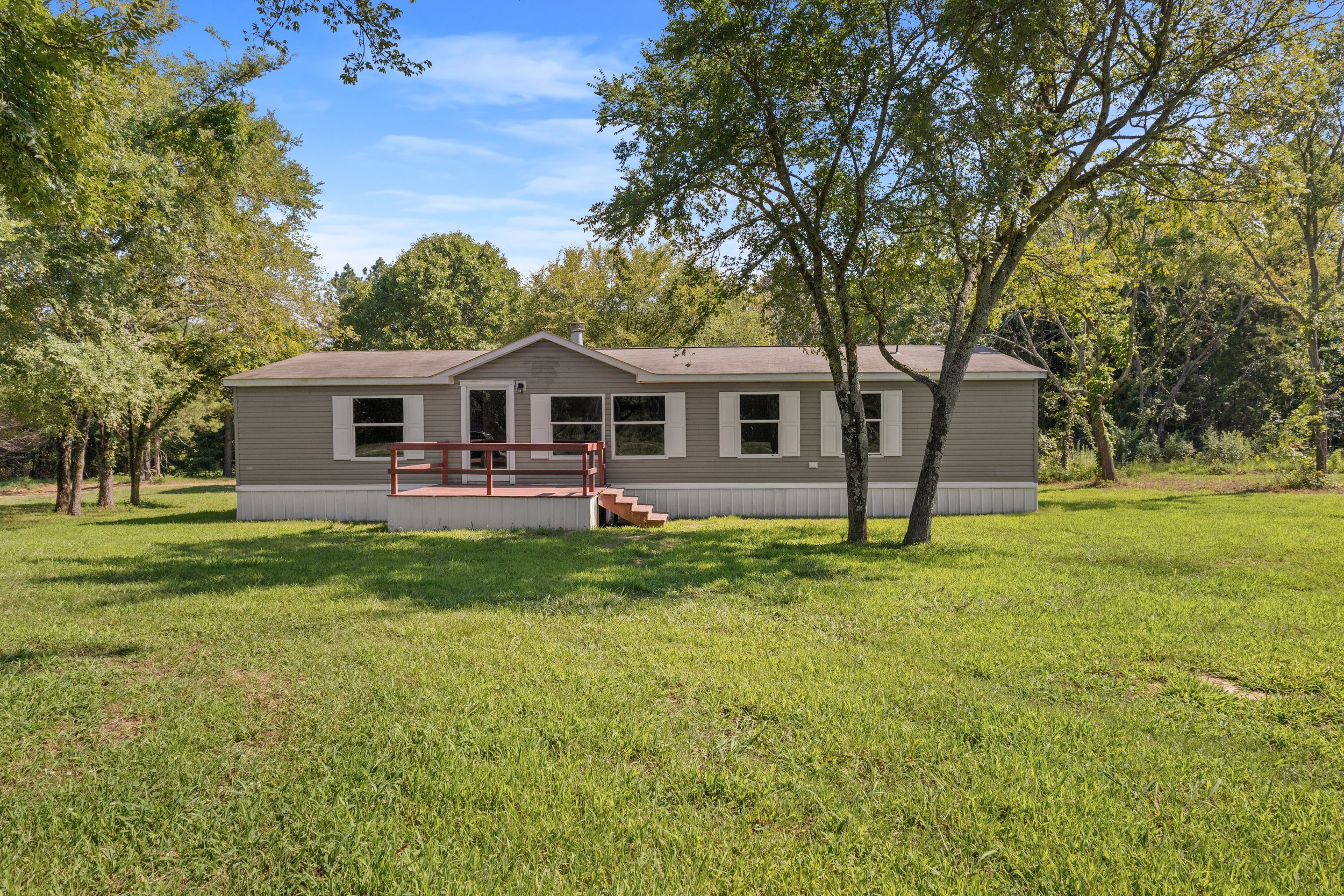 A house with beige siding, white window shutters, and a small front porch with steps, surrounded by green grass and large trees under a bright blue sky with some clouds.