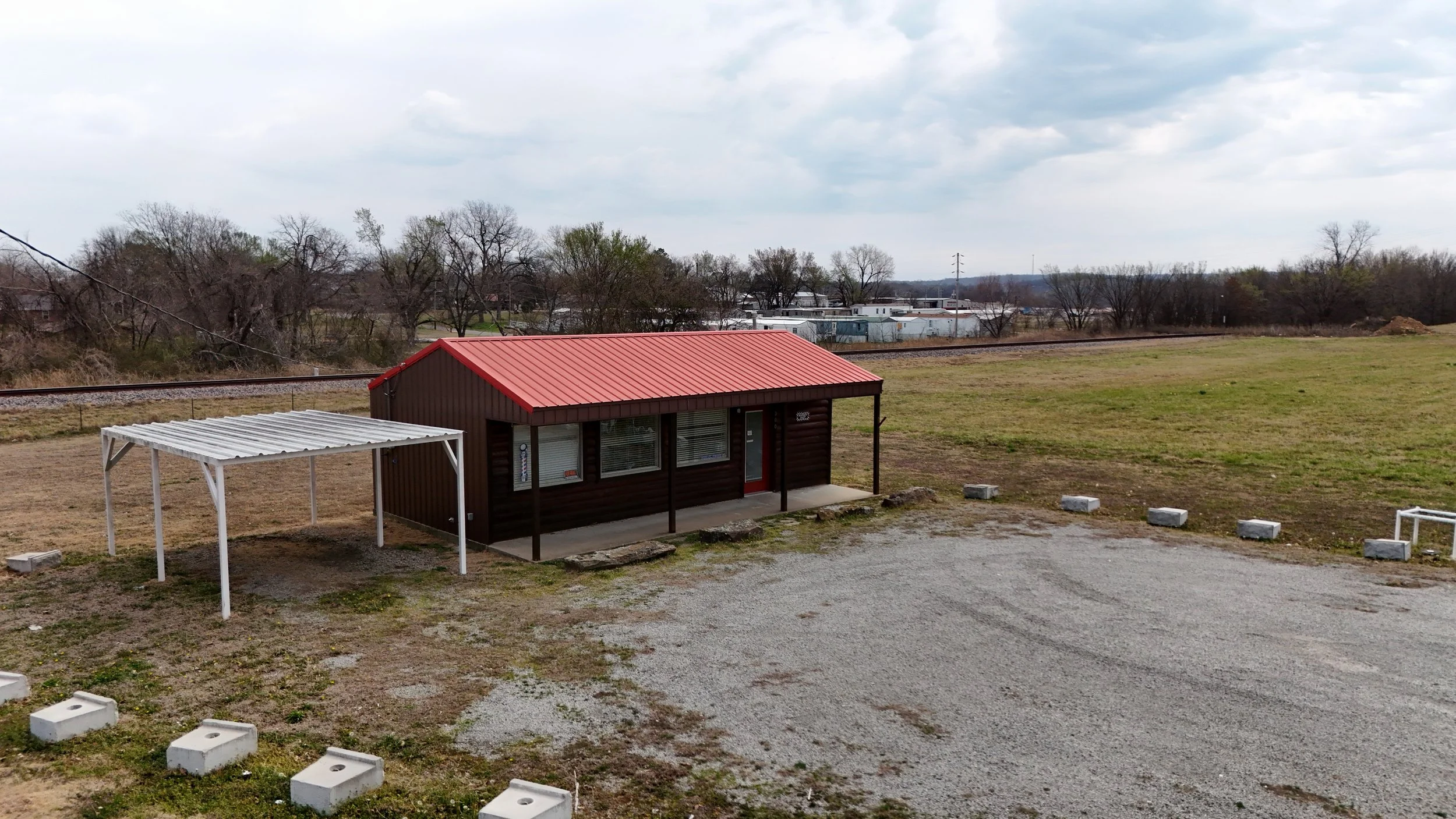 A small brown building with a red metal roof and a white covered porch, situated on a grassy area near a gravel lot, with trees and mobile homes in the background under a cloudy sky.