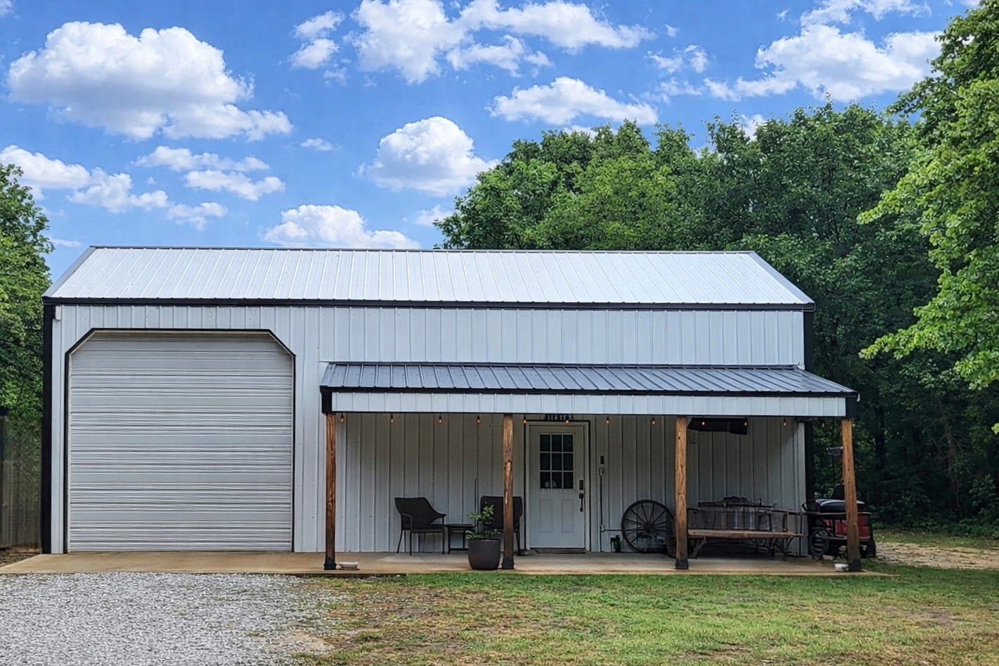 Metal barn with a large roll-up door and a smaller door under a porch, surrounded by trees and a clear sky.