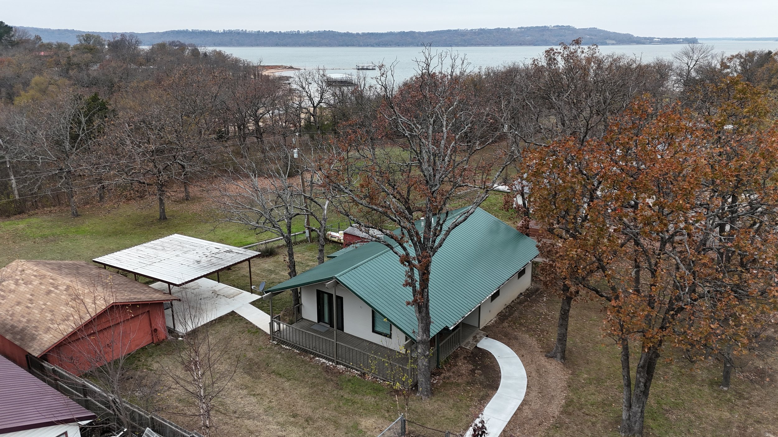 Aerial view of a backyard with a modern house, large trees with autumn leaves, a dock on a lake in the background, and overcast sky.