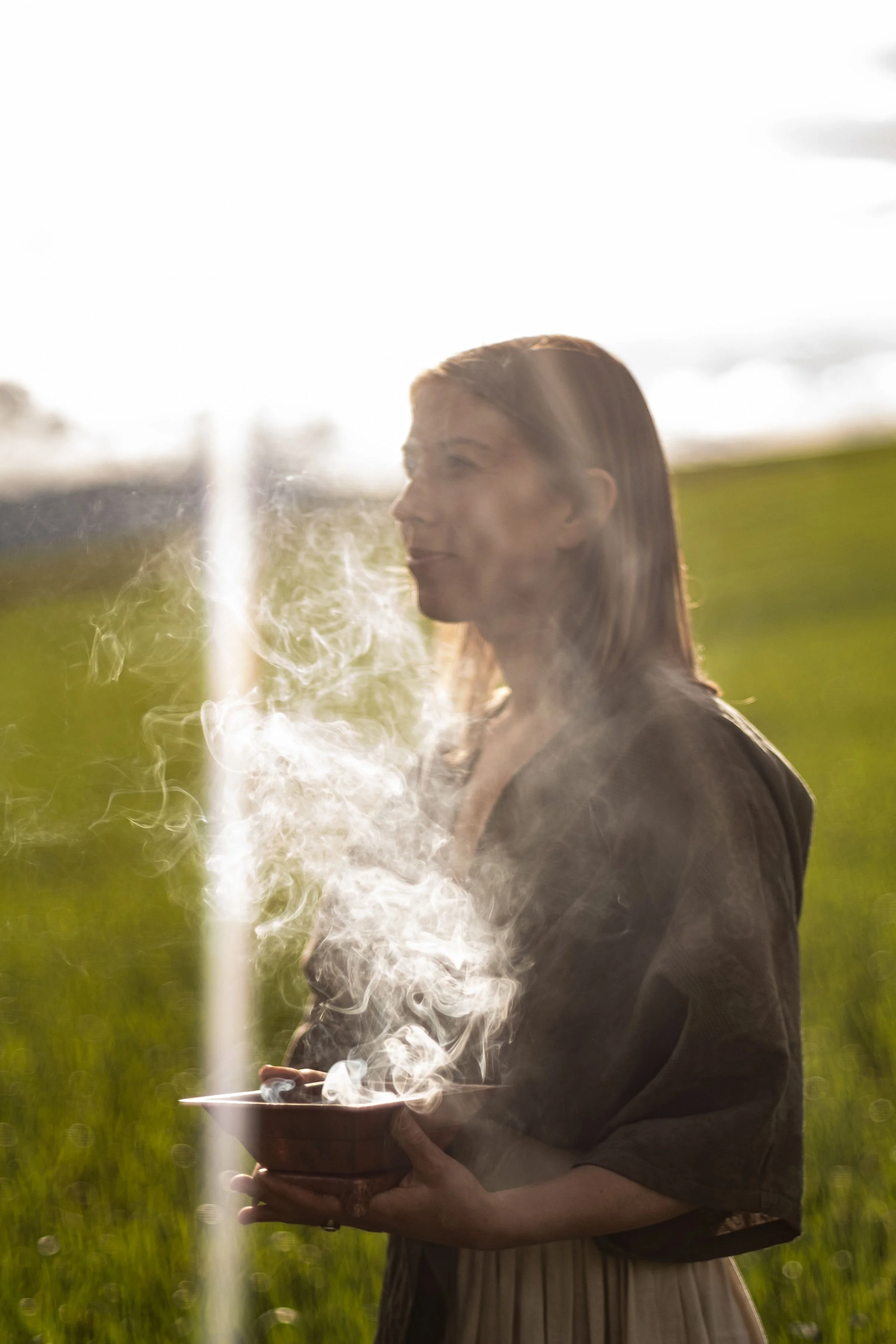 A woman standing outdoors in a field of green grass, holding a bowl with steam rising from it, backlit by the sun.