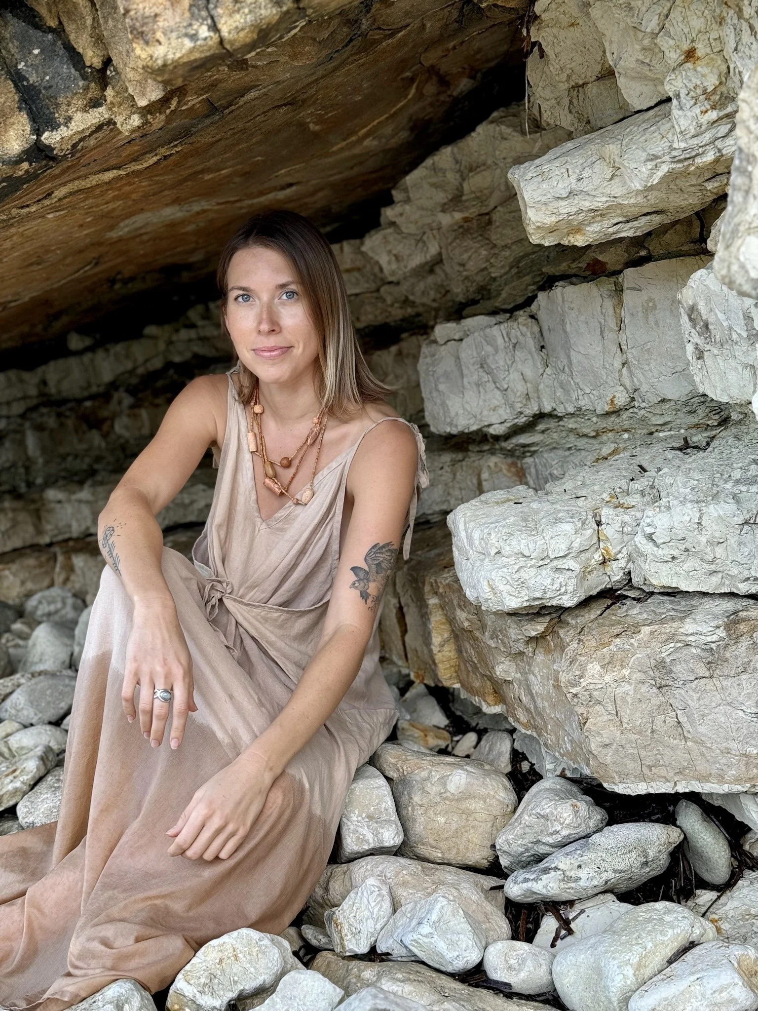A woman with shoulder-length hair, wearing a beige dress and layered necklaces, sitting on rocks with a rocky cave background.