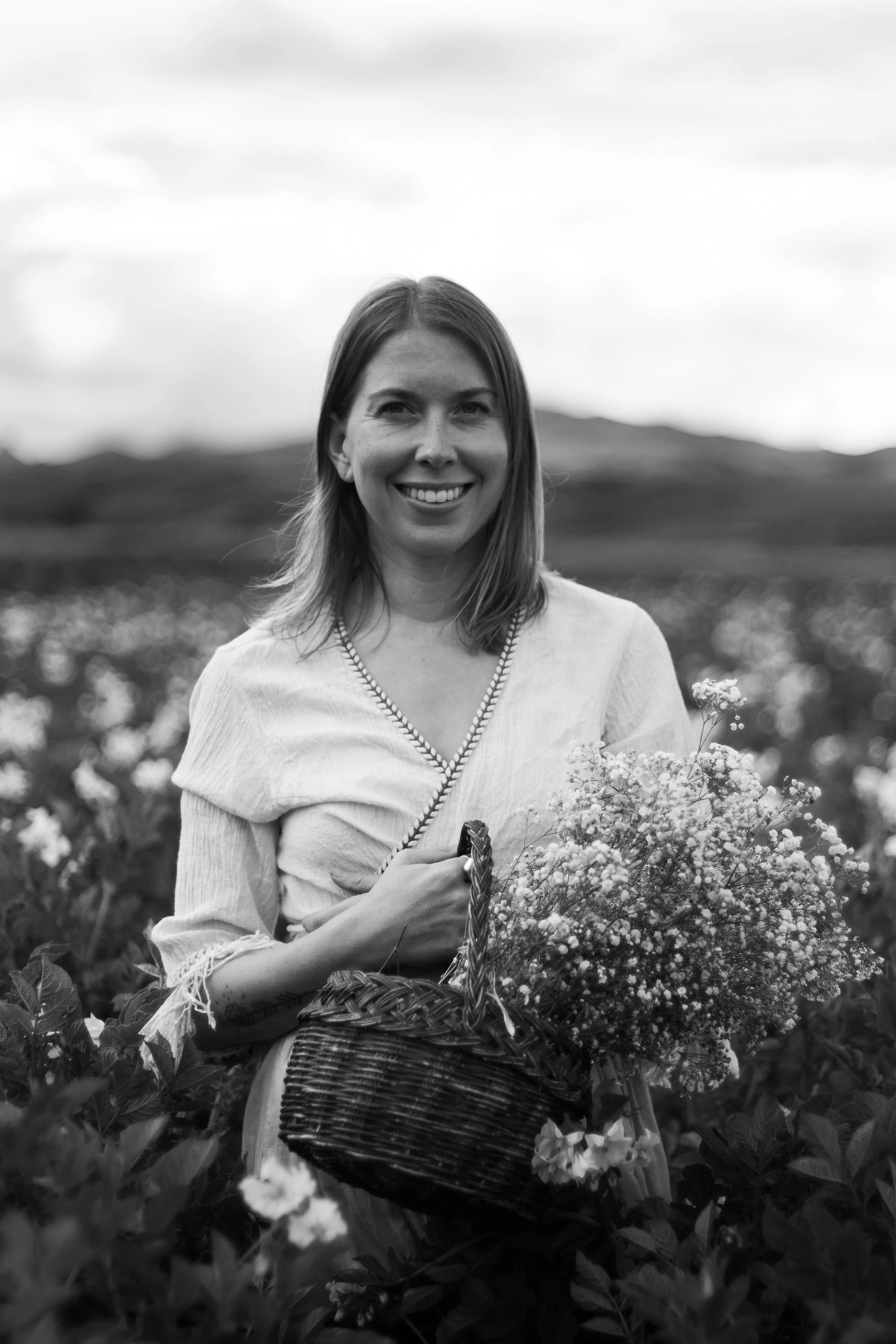 Black and white photo of a smiling woman holding a basket of flowers in a field of plants with hills in the background.