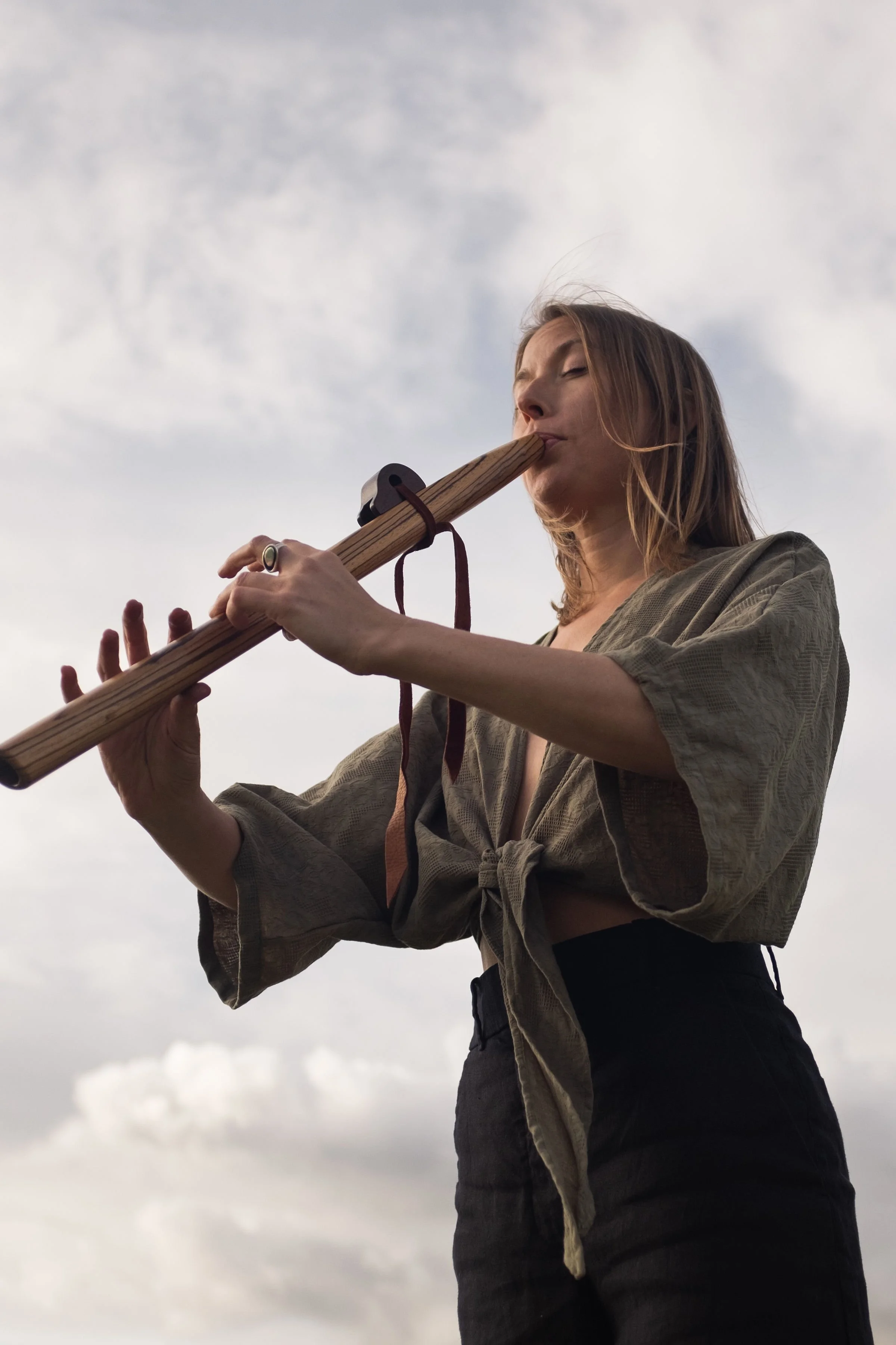 A woman playing a wooden flute outdoors against a cloudy sky.