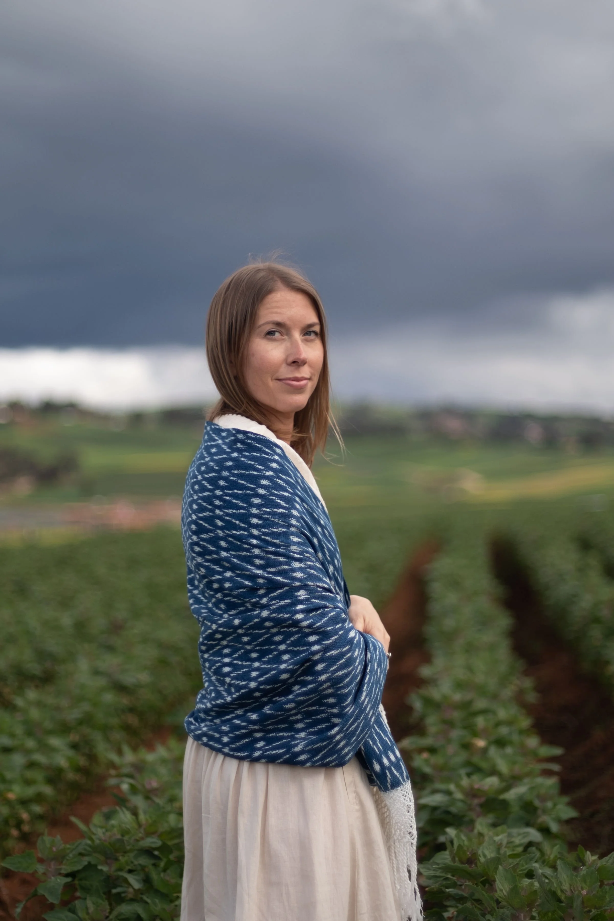 A woman standing in a farmland with rows of crops, under a cloudy, dark sky, wearing a blue patterned jacket and a light-colored skirt.