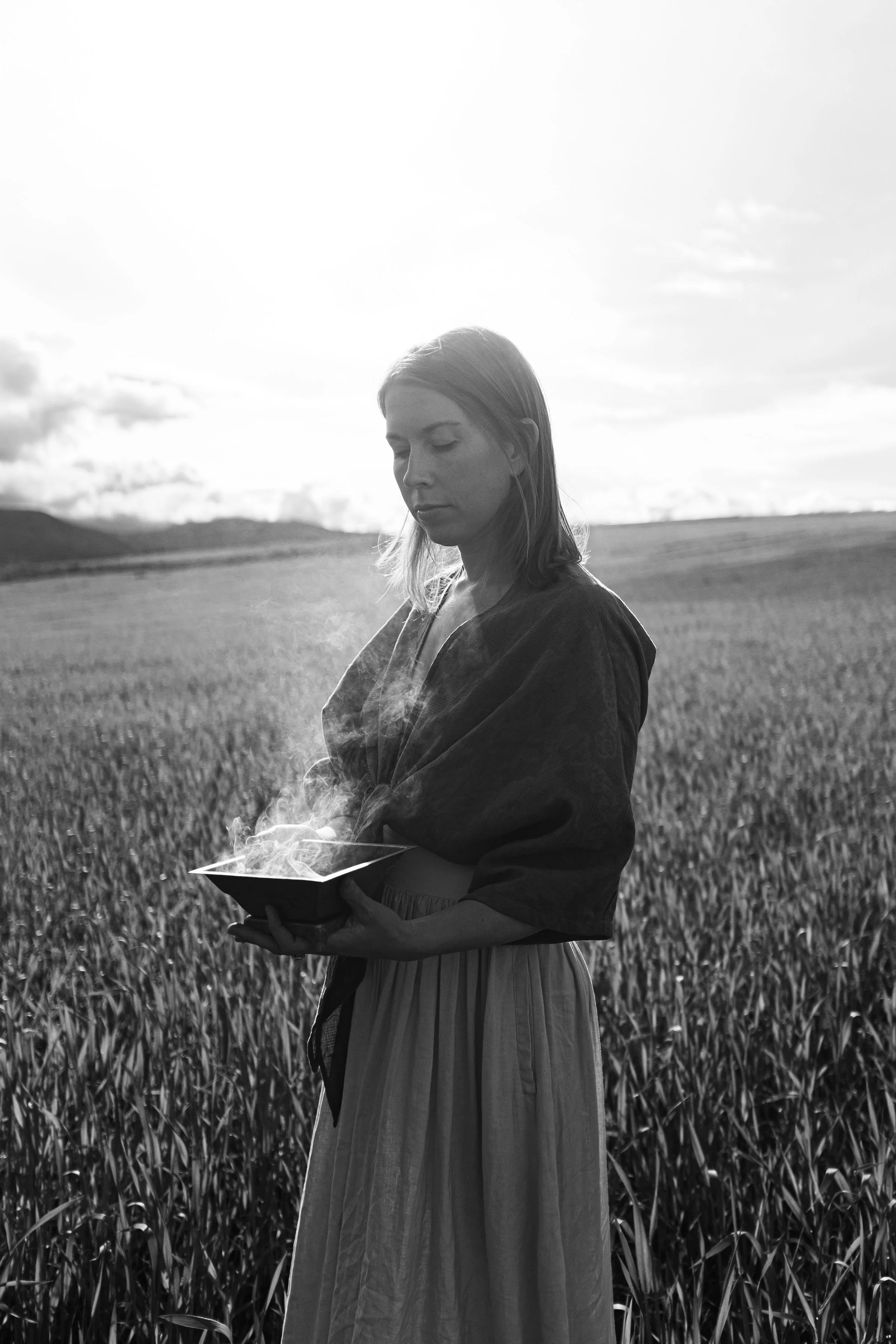 A woman in a field holding a steaming bowl, with mountains and a cloudy sky in the background, black and white photo.