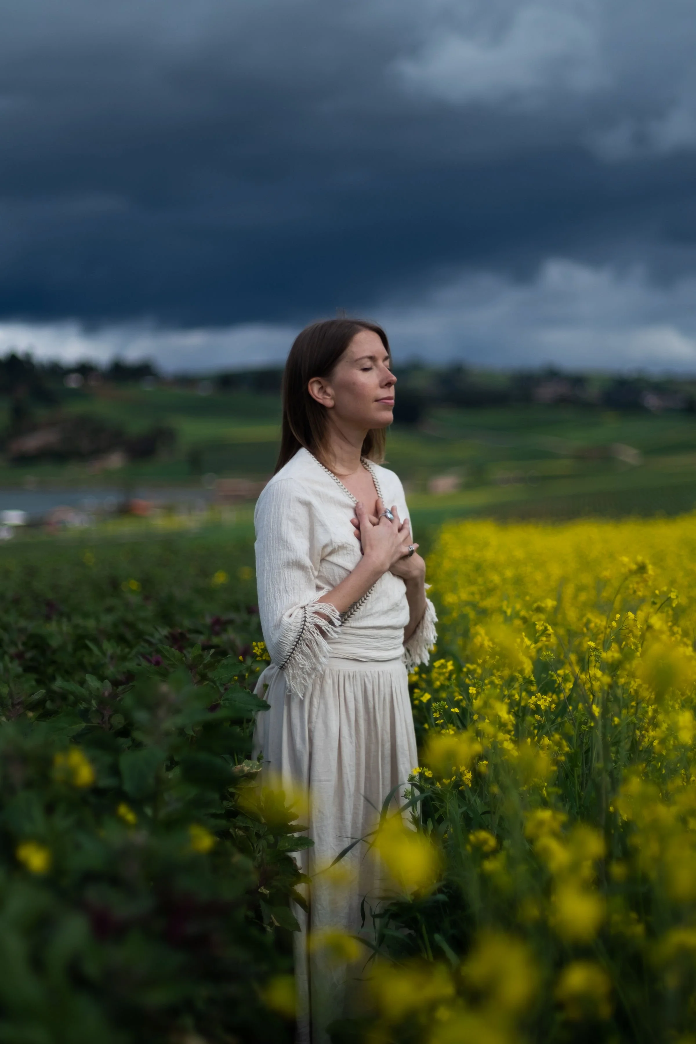 A woman with brown hair in a white dress standing in a field of yellow flowers, with dark storm clouds overhead and her hands over her chest, eyes closed.