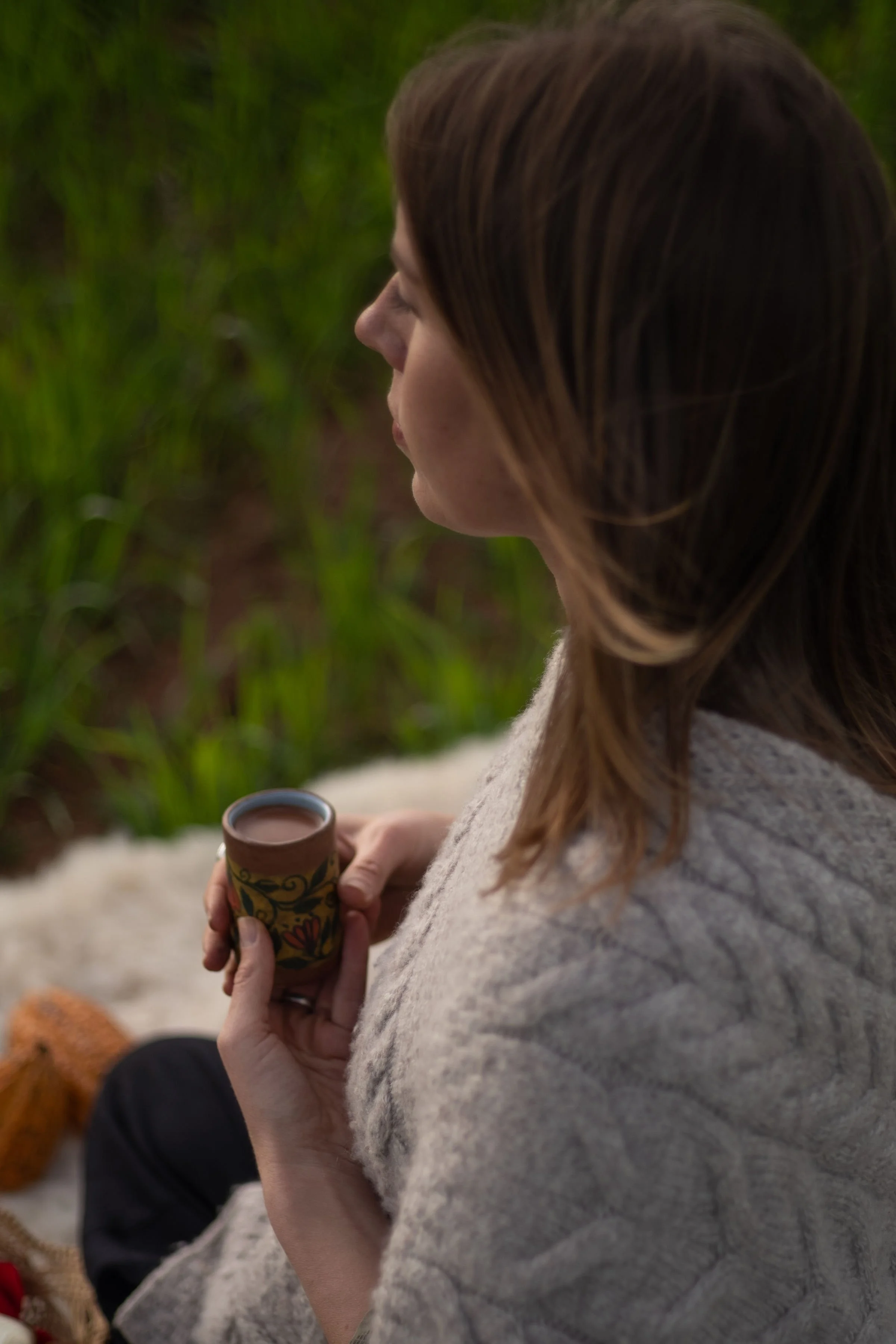 A woman with brown hair sitting outdoors holding a cup of hot beverage, with green plants in the background.