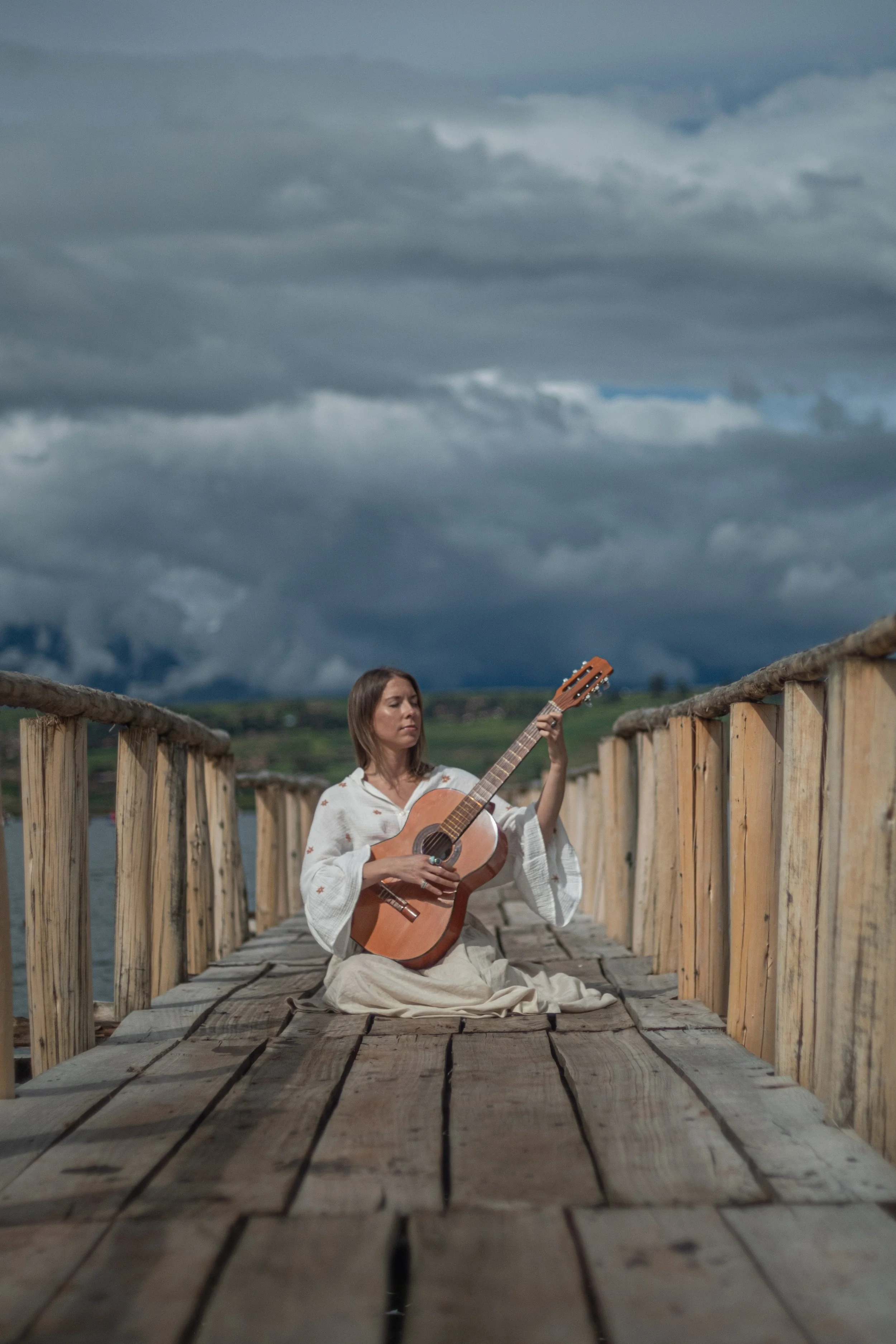 A woman in white robes sitting cross-legged on a wooden dock, playing an acoustic guitar under a cloudy sky.